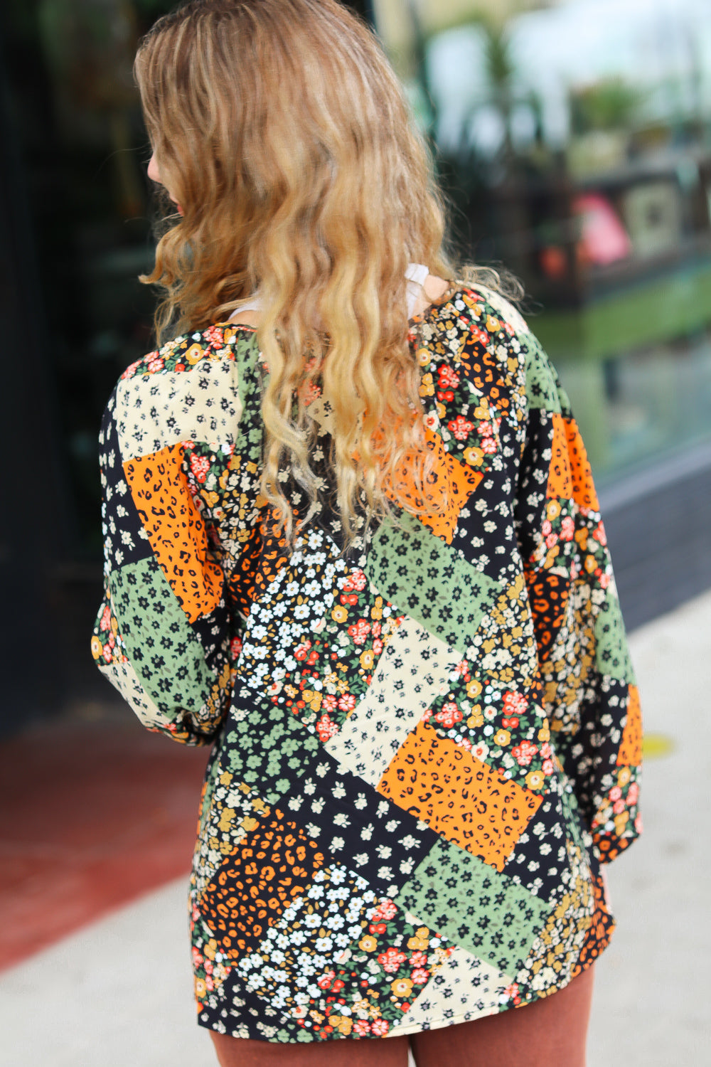 Woman with wavy blonde hair outdoors wearing a floral patchwork top with green, orange, and black accents viewed from behind