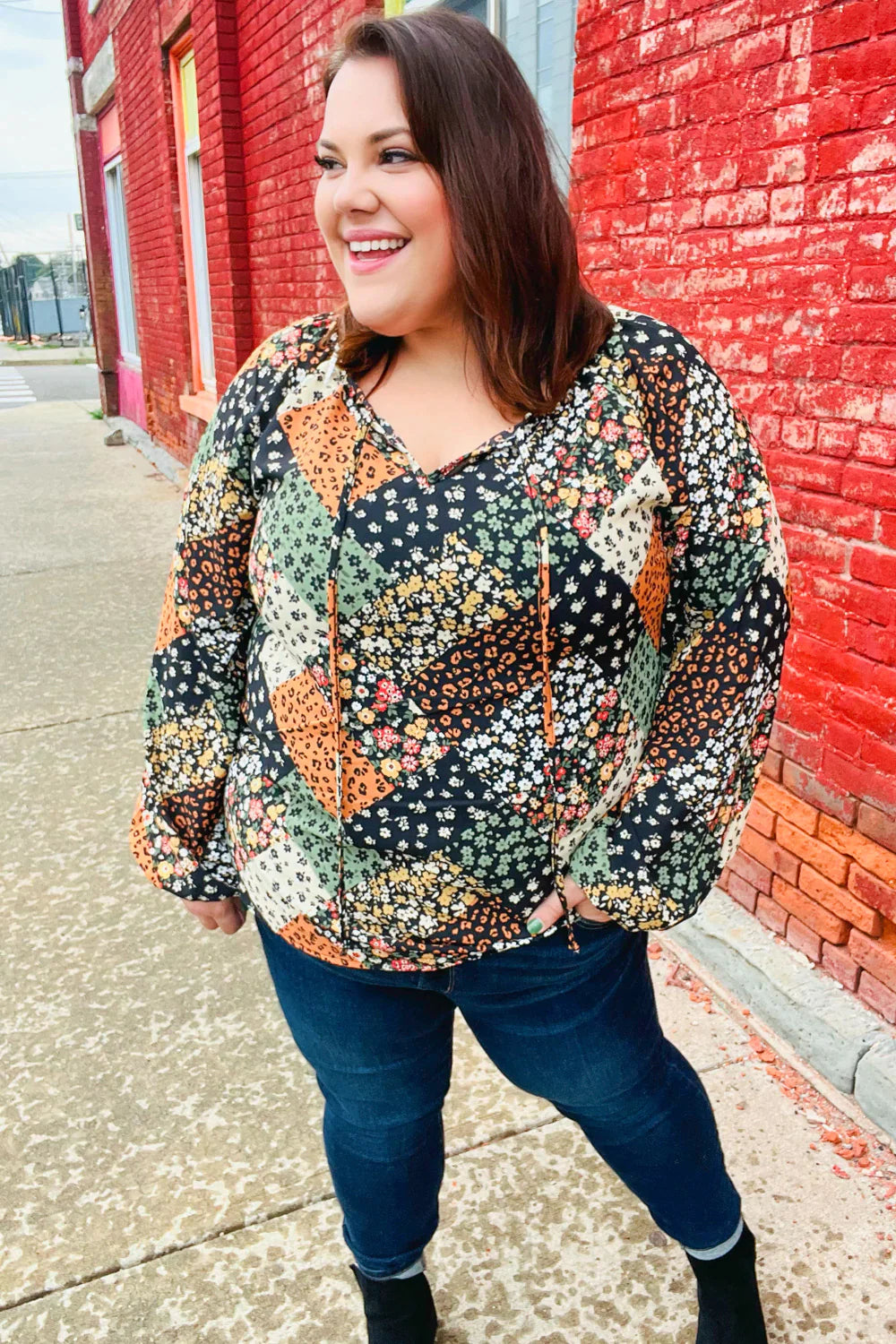 Smiling woman wearing a long sleeve floral patchwork top with keyhole neckline standing by a red brick wall outdoors