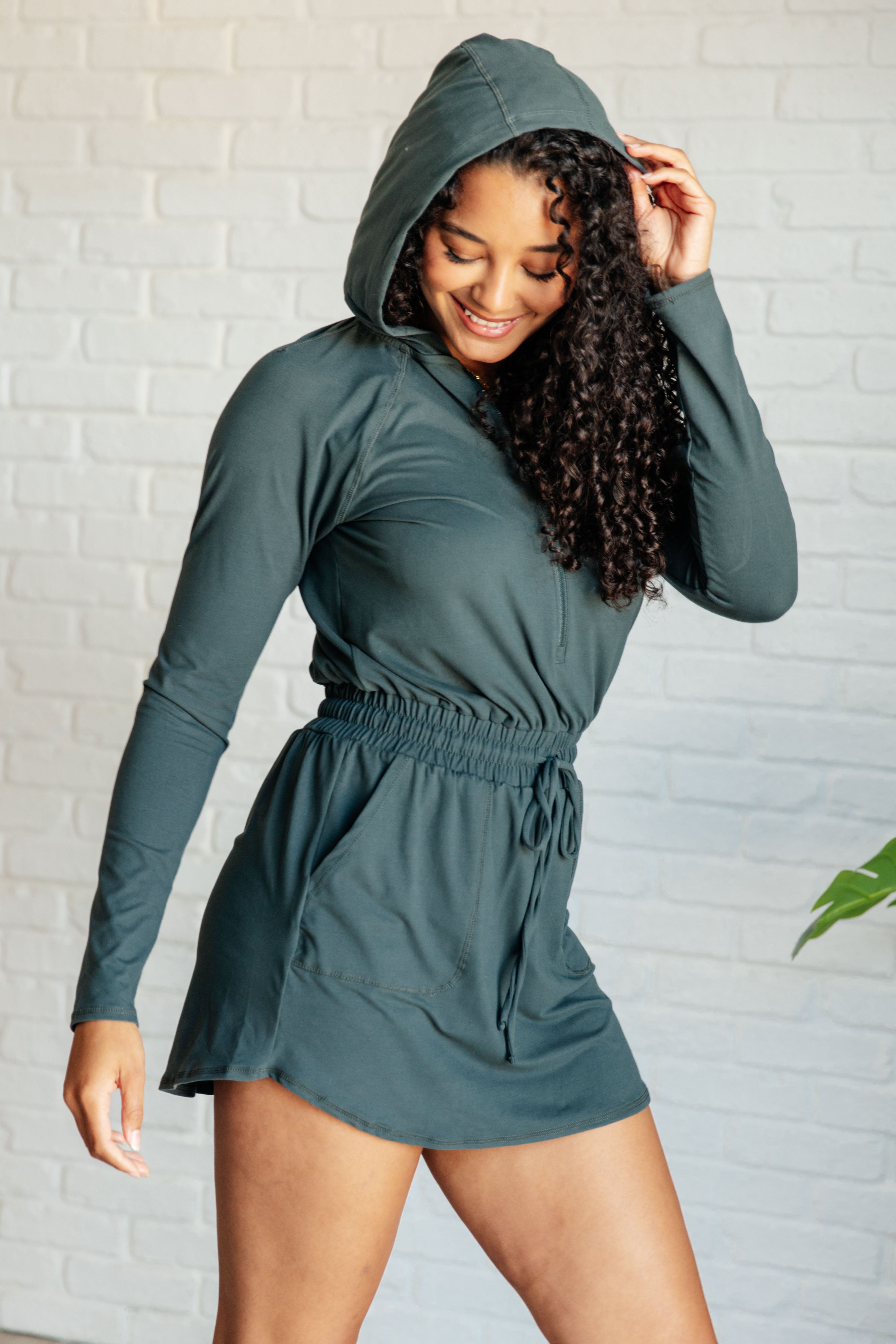 Smiling woman with curly hair wearing a smoked spruce long sleeve hoodie romper with gathered waist indoors