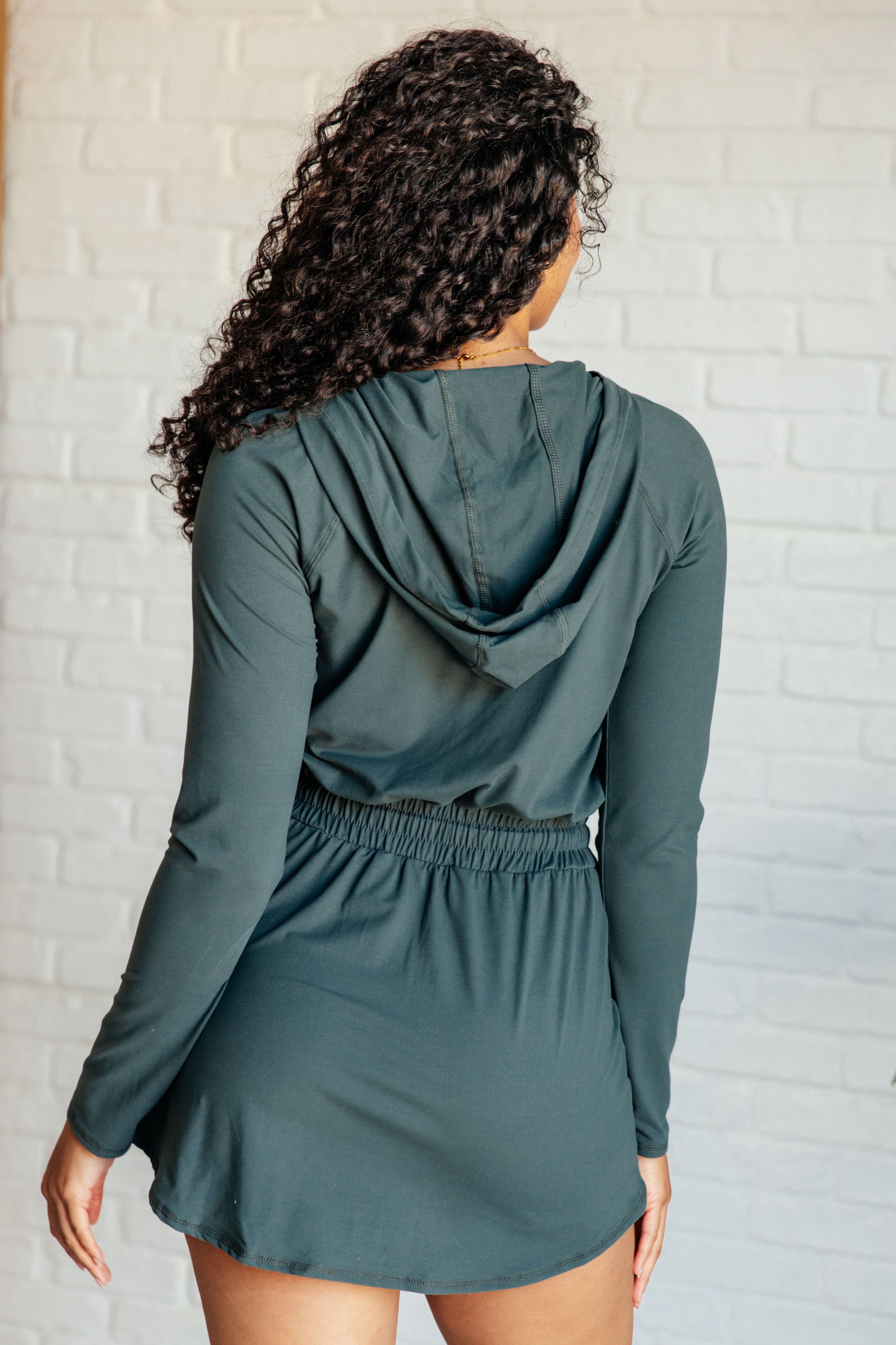 Woman with curly hair seen from behind wearing a smoked spruce long sleeve hoodie romper indoors