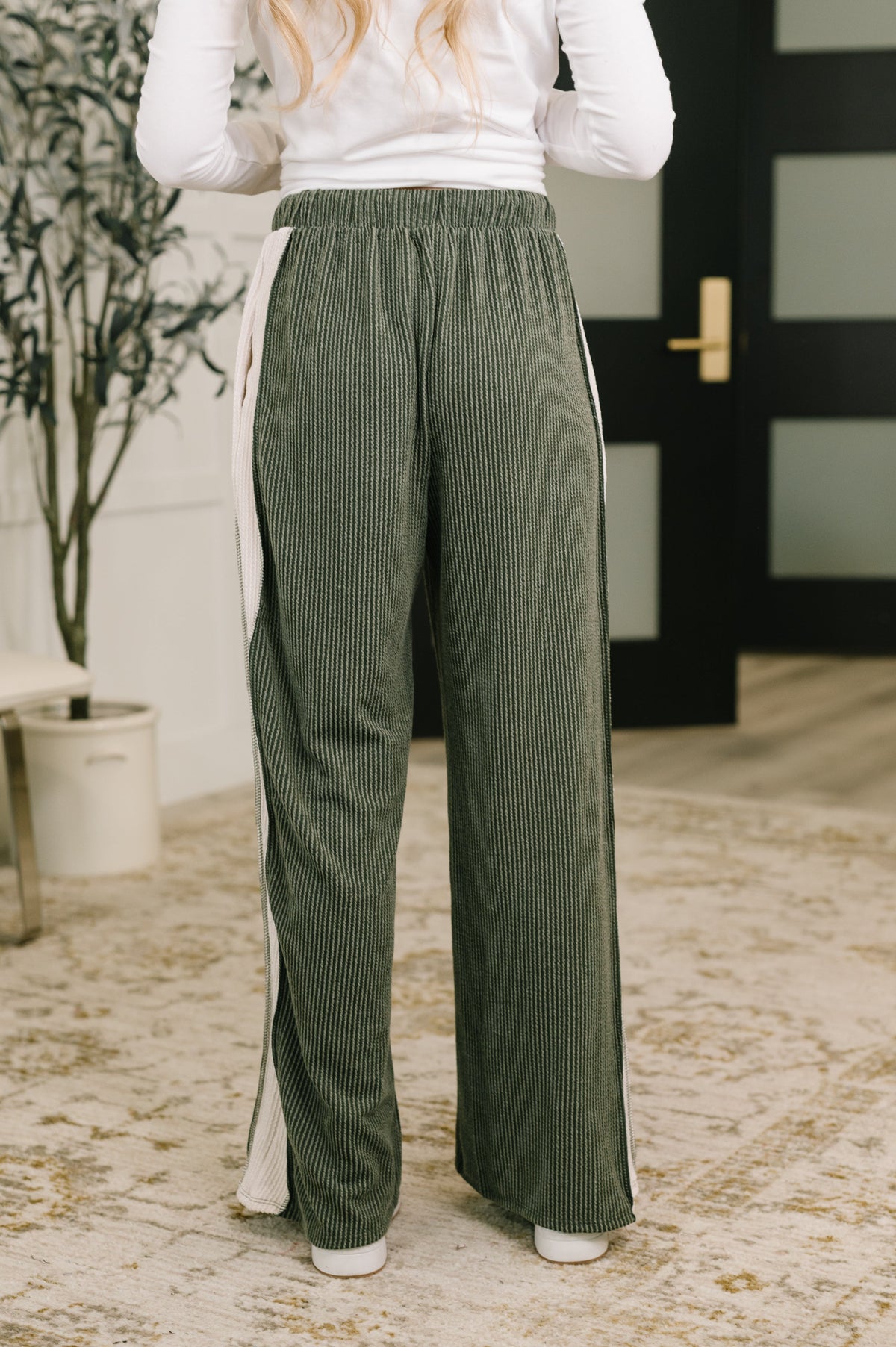 Woman wearing olive high waisted ribbed knit pants with white side stripes and wide legs while standing indoors barefoot