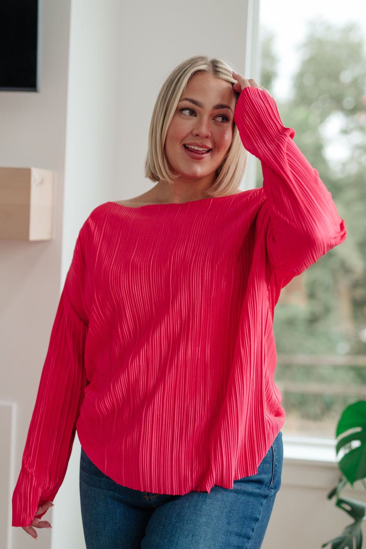 Woman wearing a fuchsia accordion pleated top with long sleeves and a relaxed neckline styled with jeans in a bright home interior