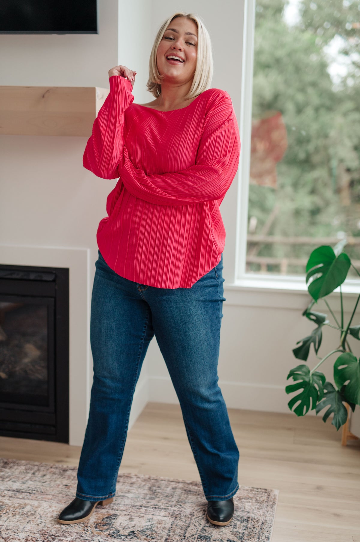 Woman wearing a fuchsia accordion pleated satin top with long sleeves and jeans while posing in a bright living room