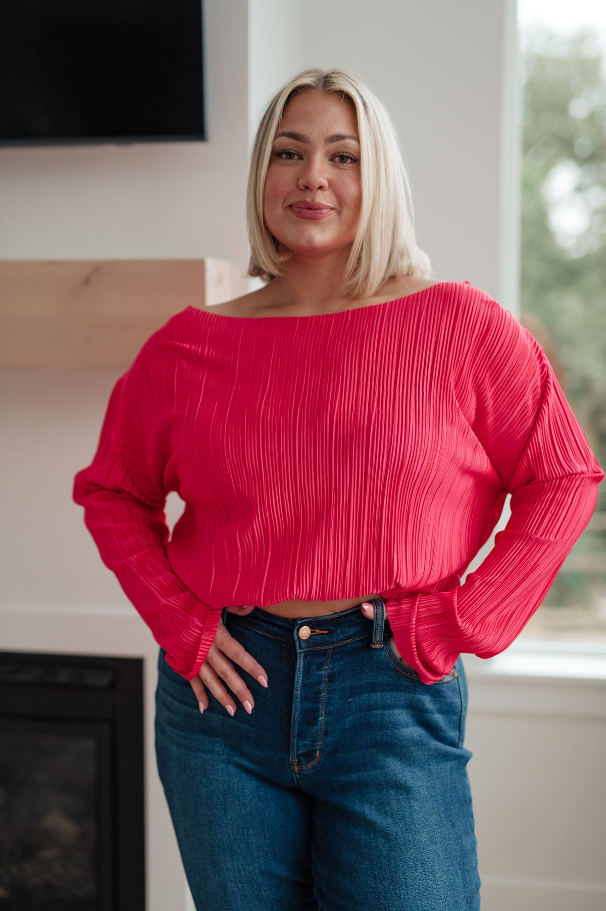 Woman wearing a fuchsia accordion pleated top with a wide neckline and long sleeves styled with blue jeans indoors