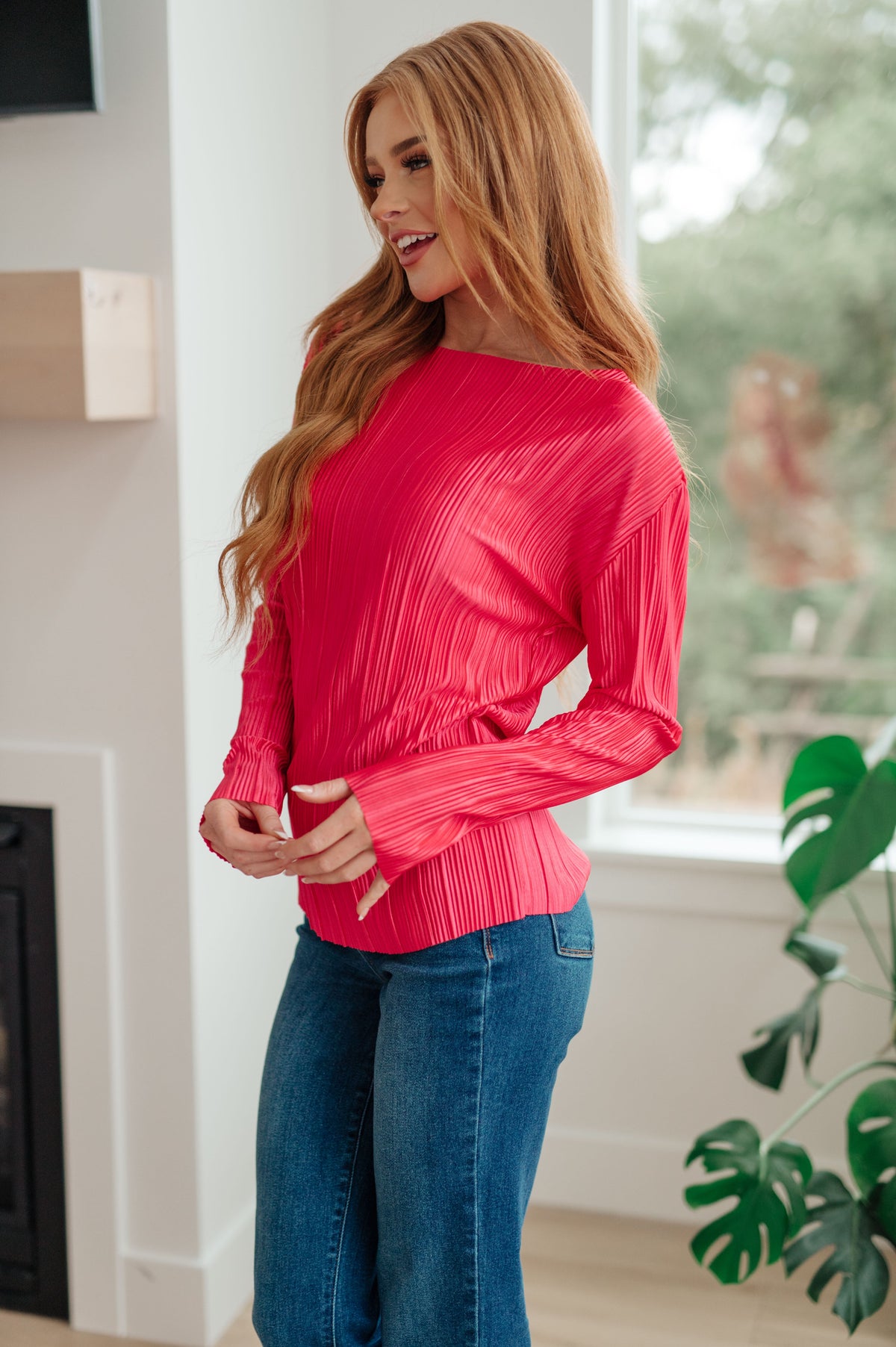 Woman wearing a vibrant fuchsia pleated satin top with long sleeves styled with blue jeans in a bright home interior