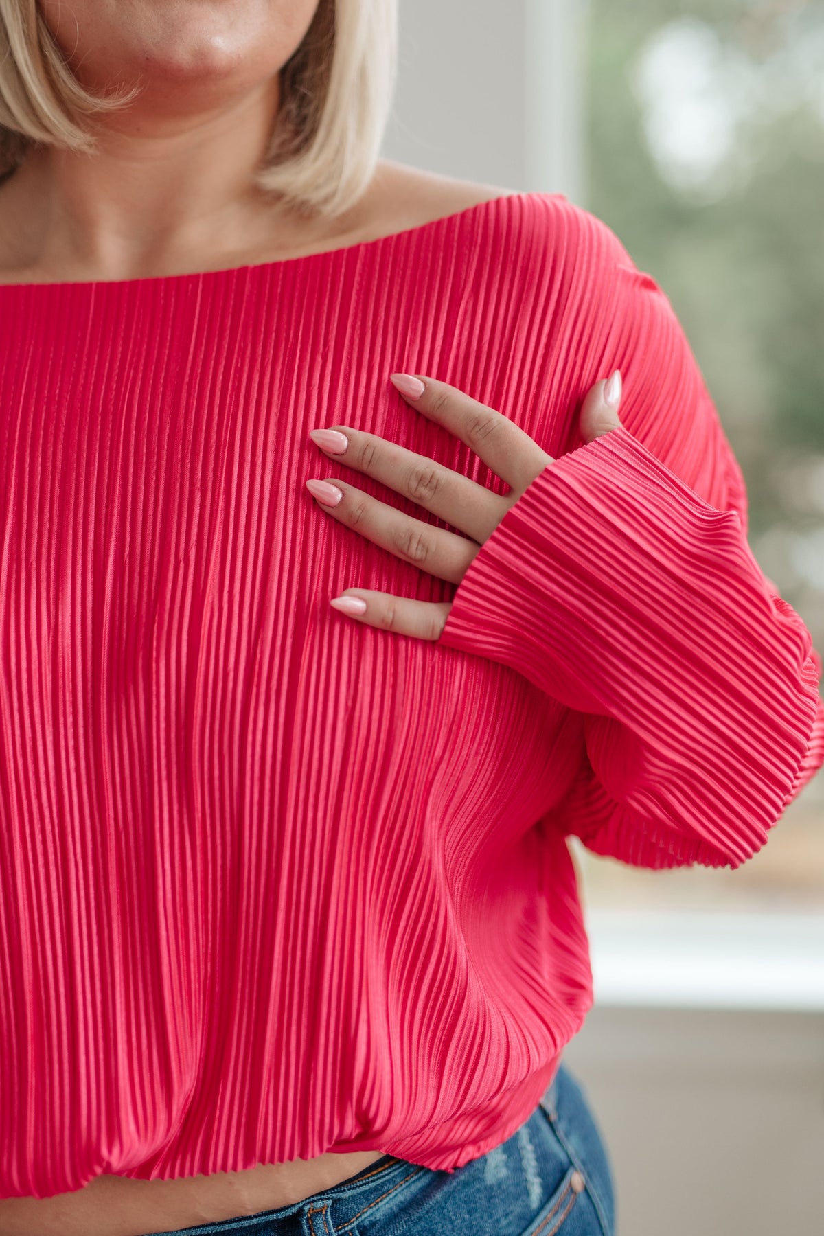 Close view of a woman wearing a fuchsia accordion pleated satin top with a wide neckline and flared sleeves styled with jeans indoors