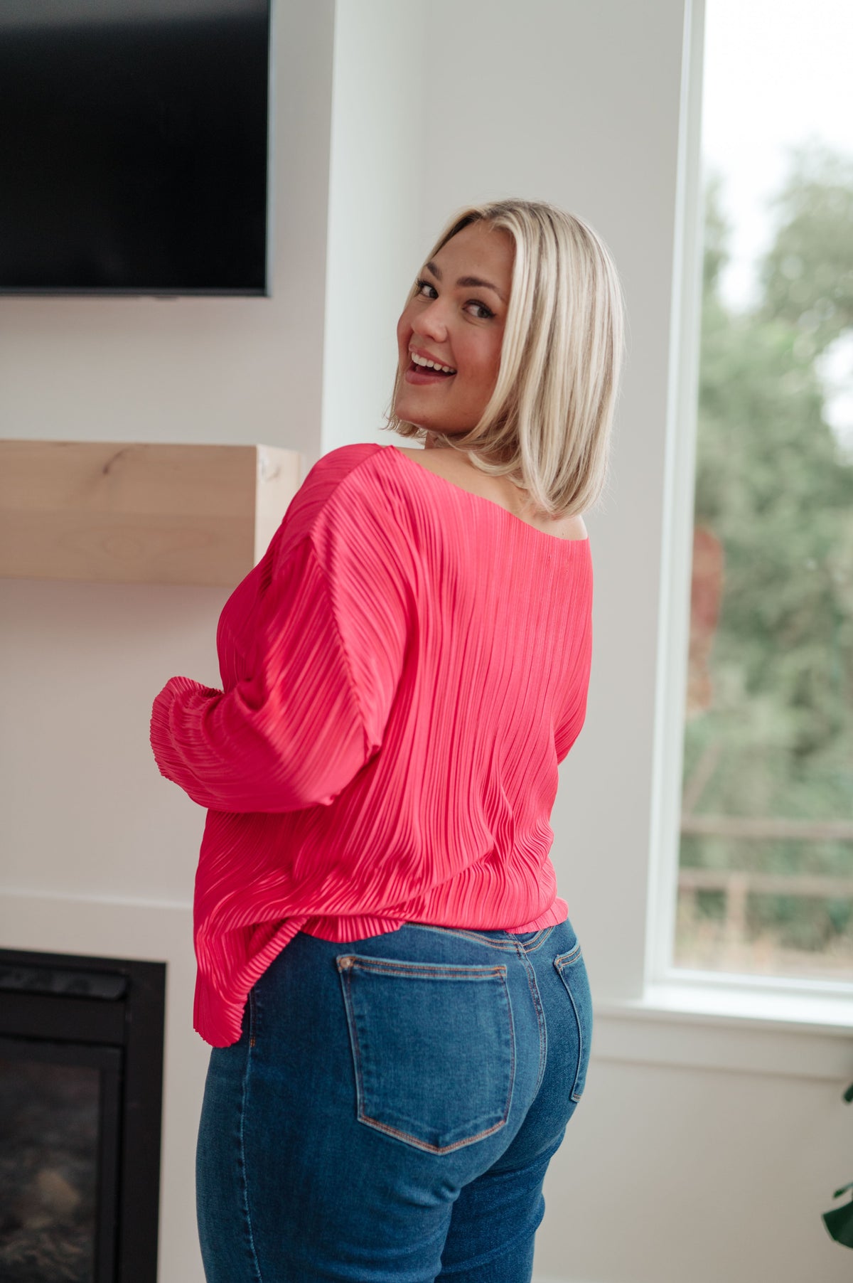 Woman looking back in a bright fuchsia pleated satin top with billowy sleeves and blue jeans beside a window