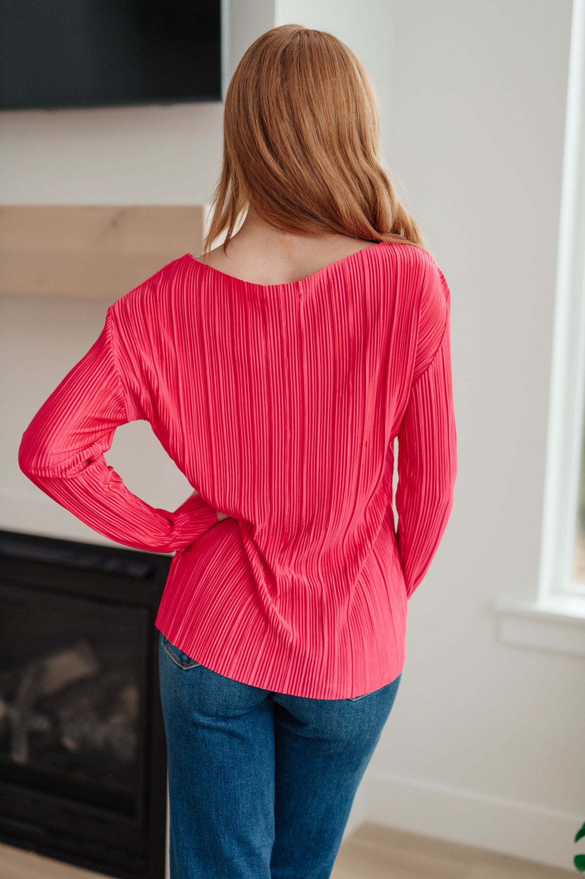 Back view of a woman wearing a vibrant fuchsia accordion pleated top with long sleeves paired with blue jeans in a bright room