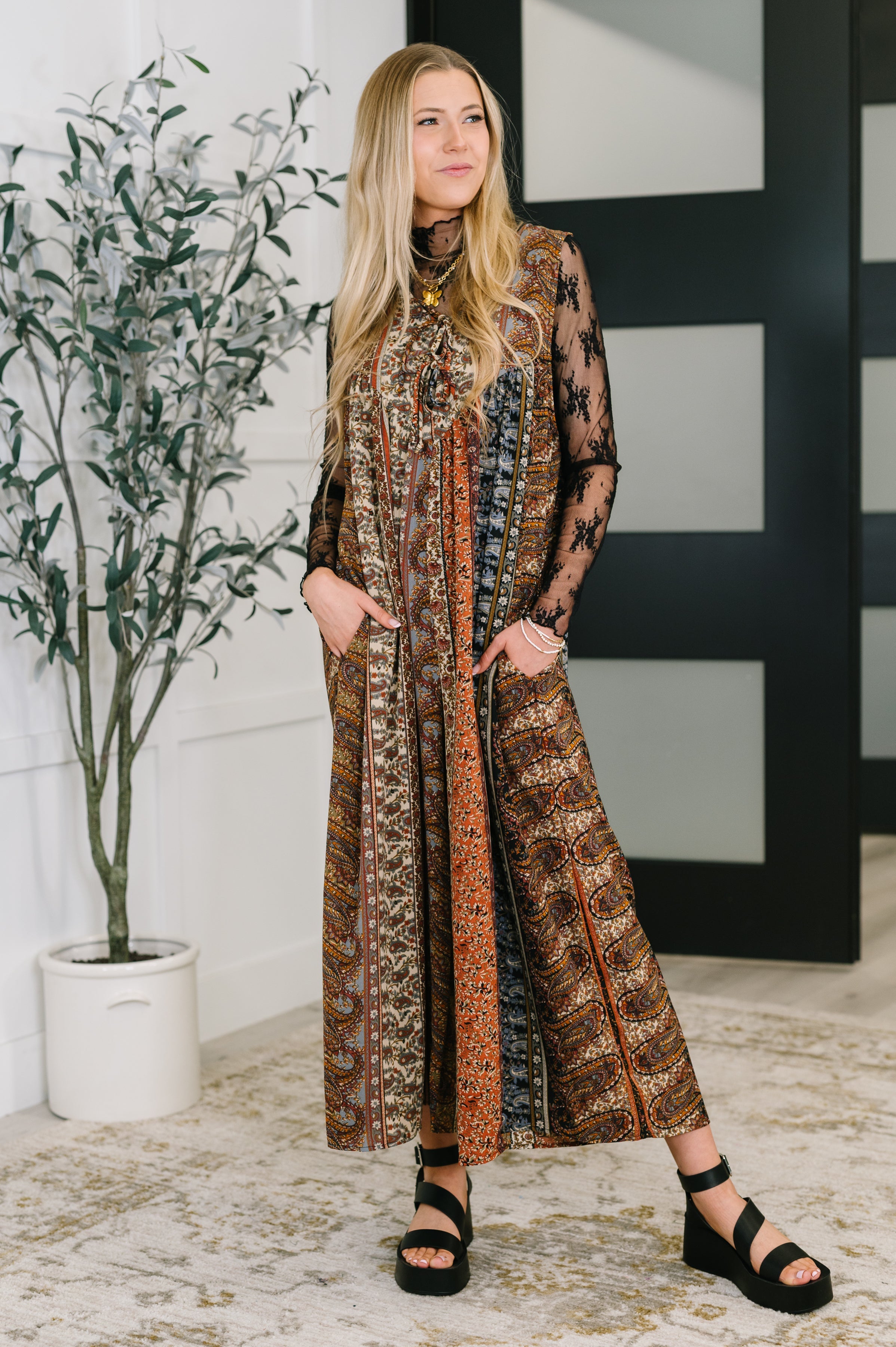 Blonde woman wearing a brown mixed print jumpsuit with long sheer black floral sleeves standing indoors near a plant
