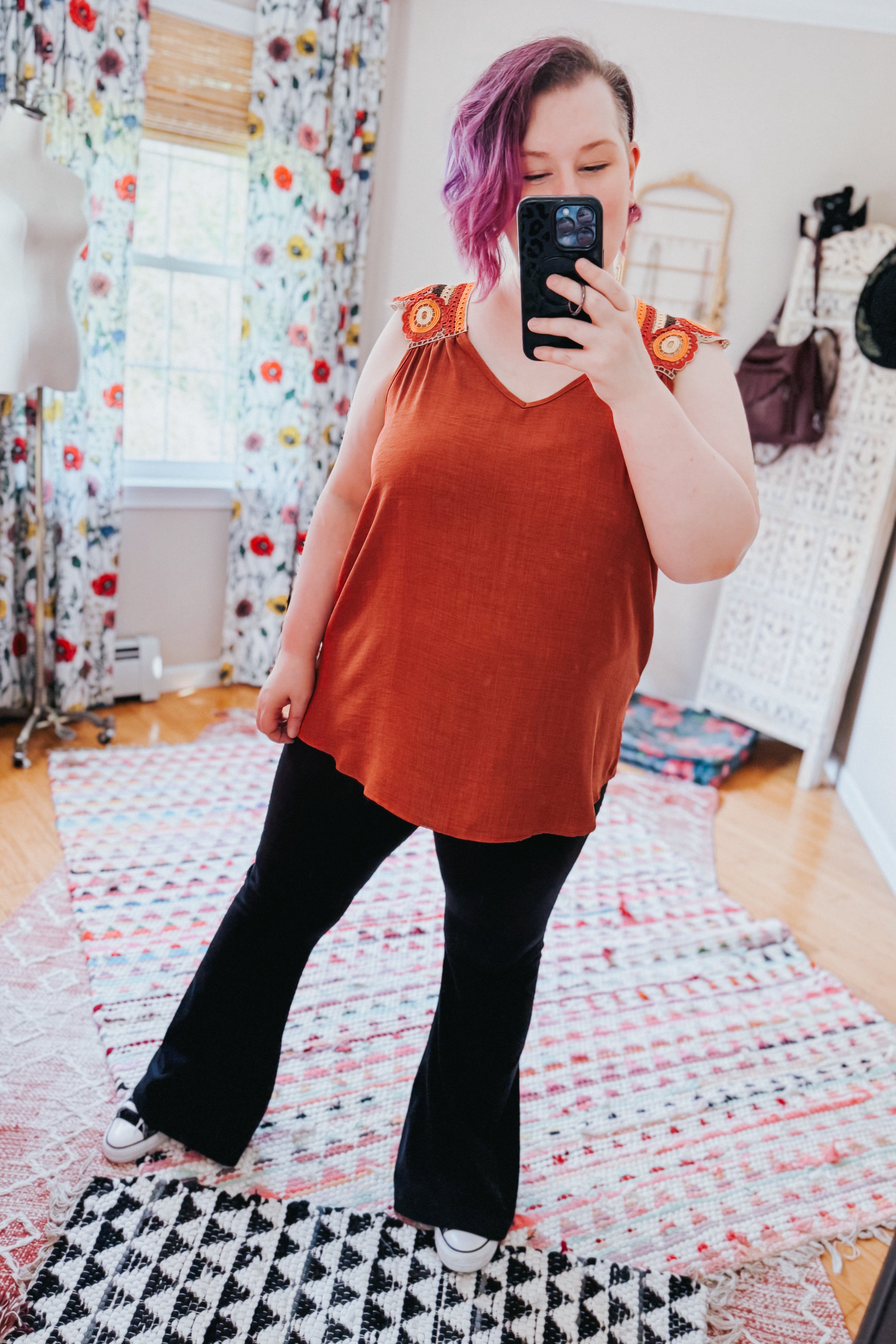 Woman wearing an orange sleeveless top with crochet floral shoulder details and black flared pants taking a mirror selfie