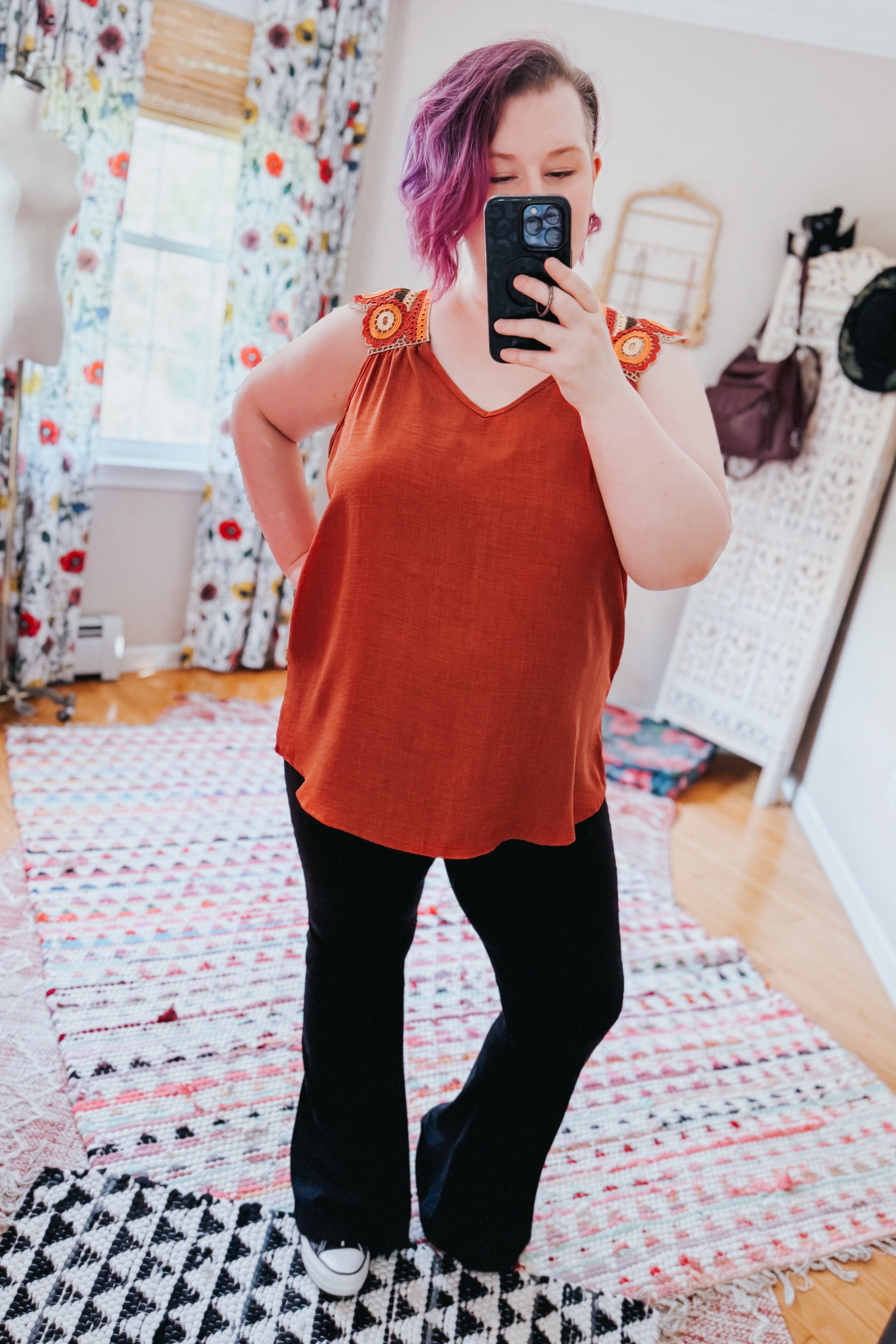 Woman taking mirror selfie wearing a burnt orange sleeveless top with crochet floral shoulder accents and black pants