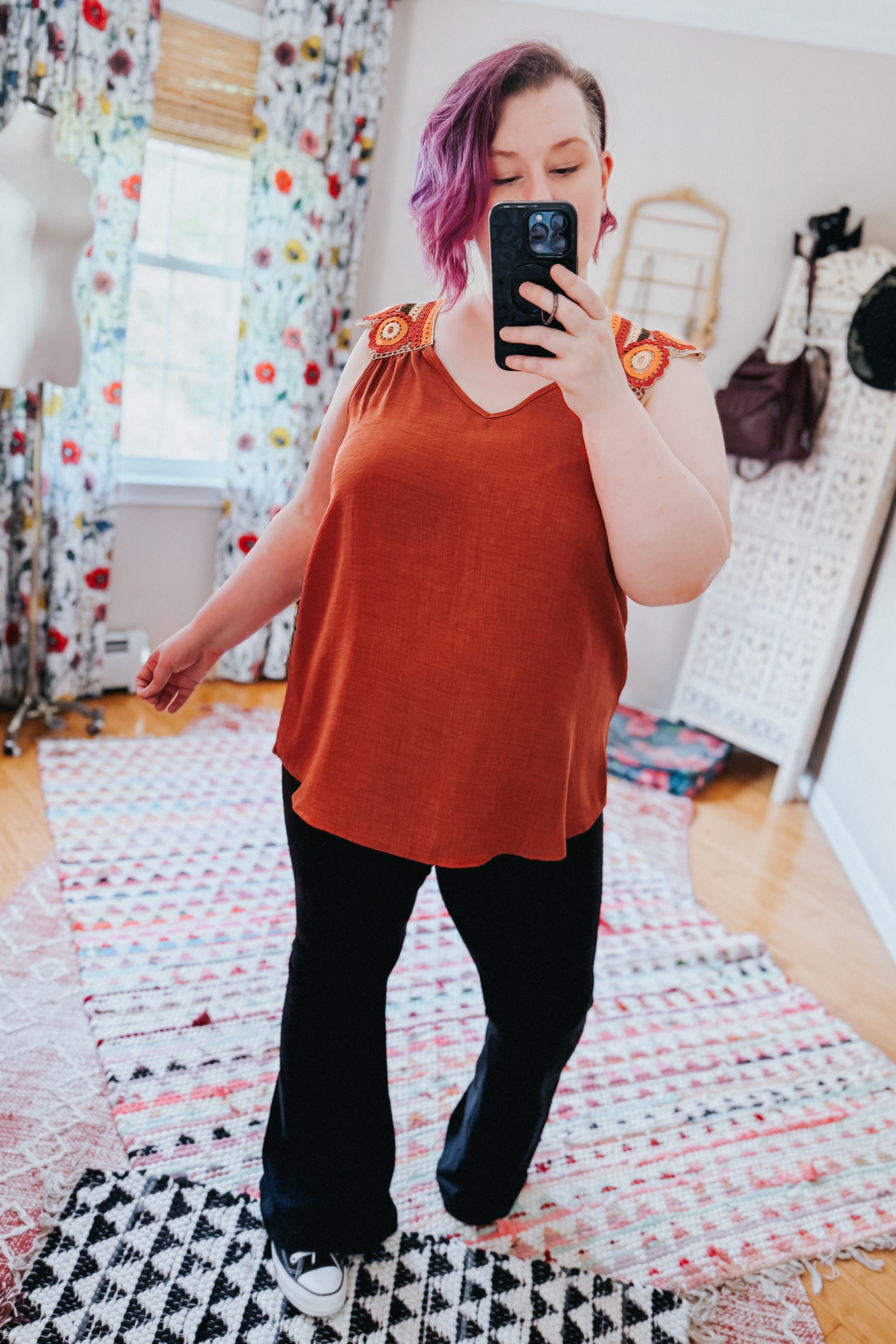 Woman taking mirror selfie wearing a rust orange sleeveless top with crochet shoulder details and black pants