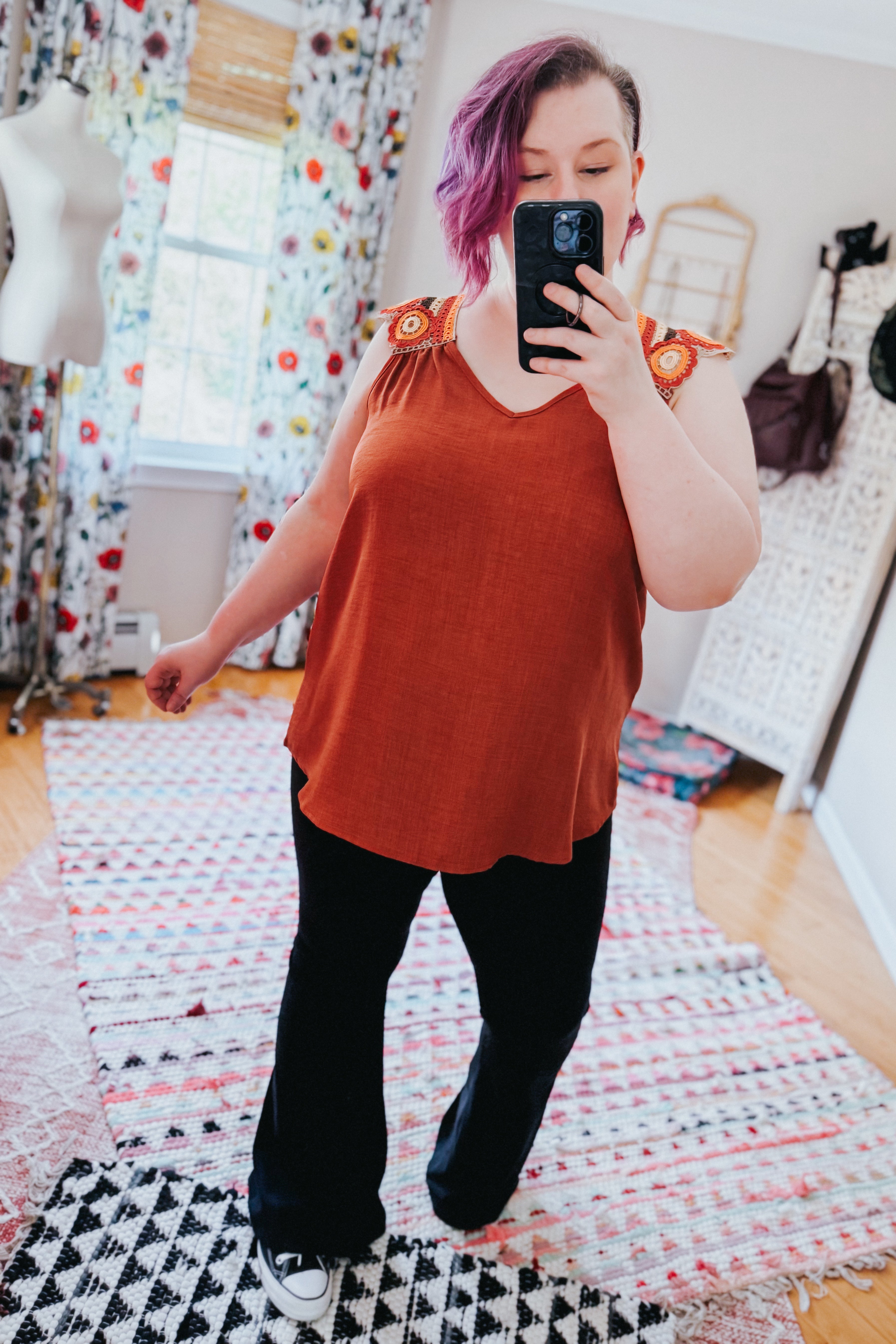 Woman taking mirror selfie wearing an orange sleeveless top with crochet floral shoulder embellishments and black pants