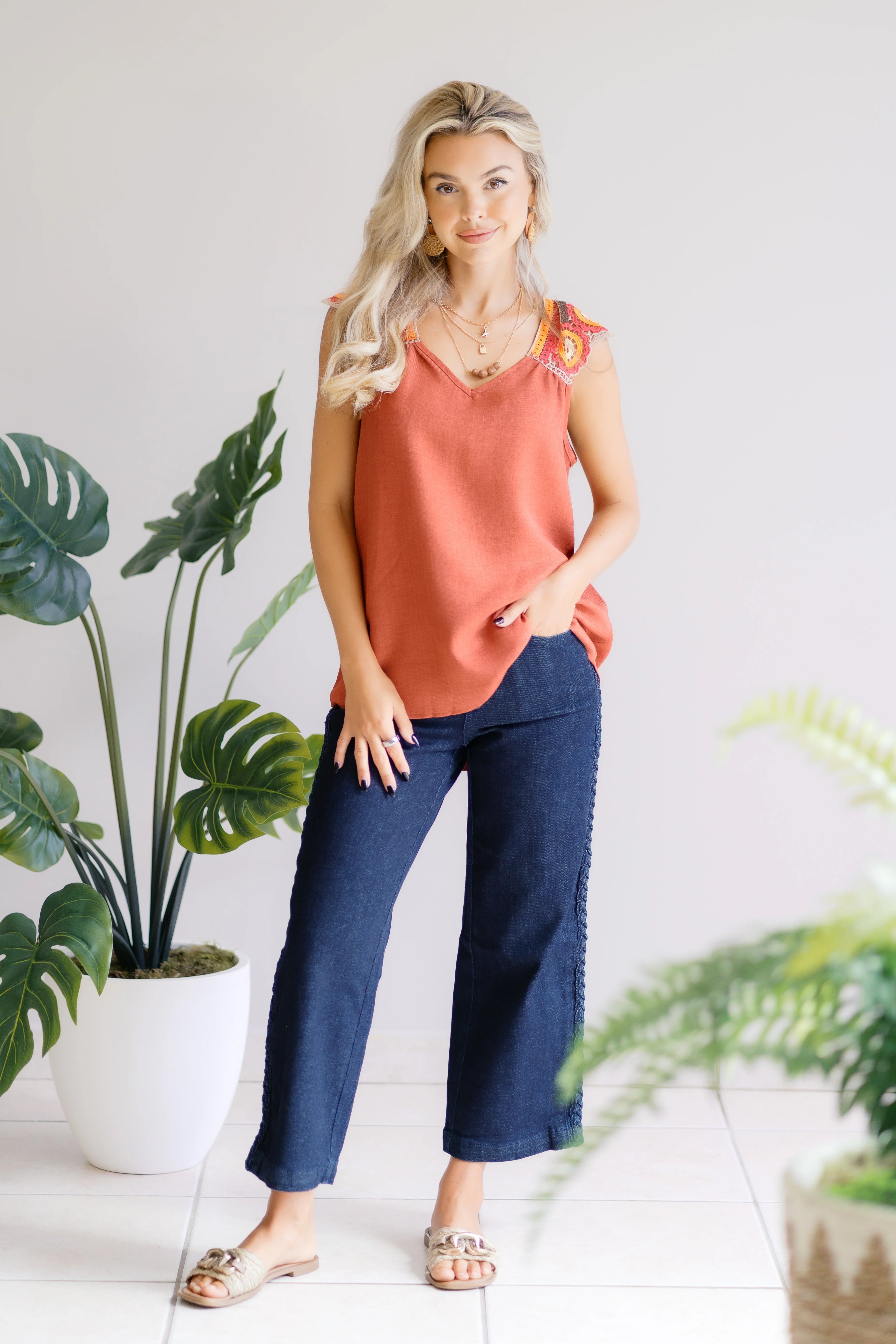 Blonde woman wearing an orange sleeveless top with crochet floral shoulder details and wide-leg dark blue pants indoors