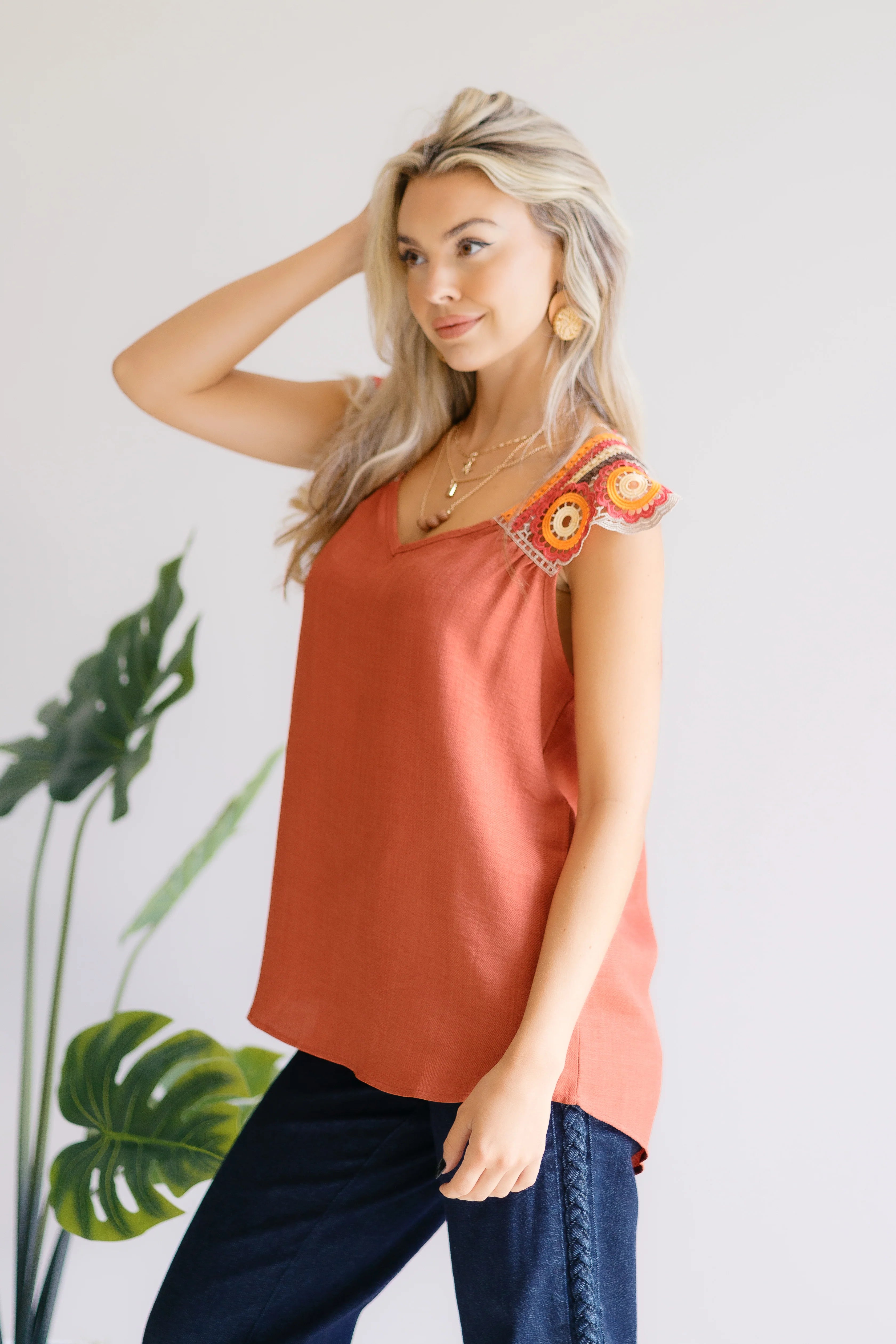 Blonde woman wearing a rust orange sleeveless top with colorful crochet floral shoulder accents and dark pants indoors