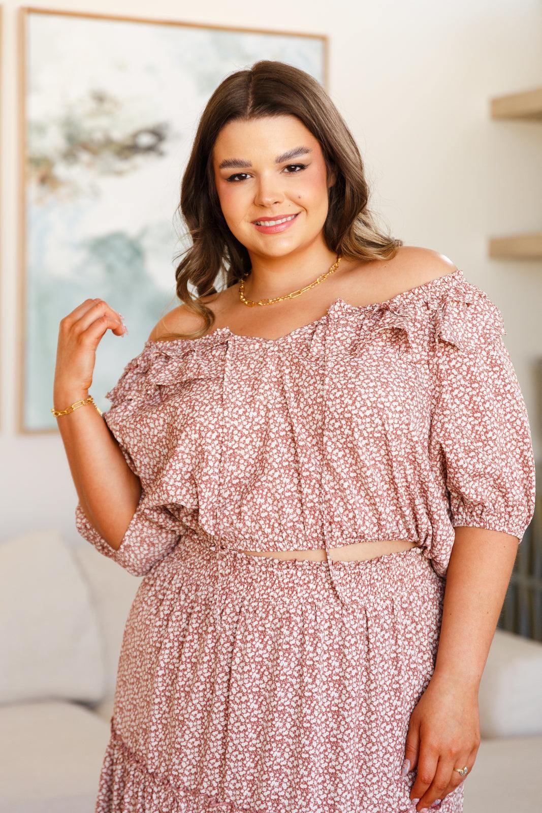 Plus size woman wearing an off-shoulder rose floral top with ruffle details and matching skirt indoors smiling