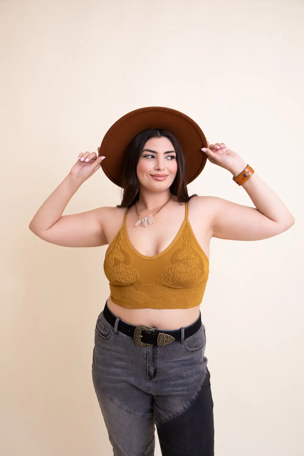 Woman adjusting wide-brim brown hat wearing mustard textured seamless brami and black and gray high-waisted jeans against neutral wall
