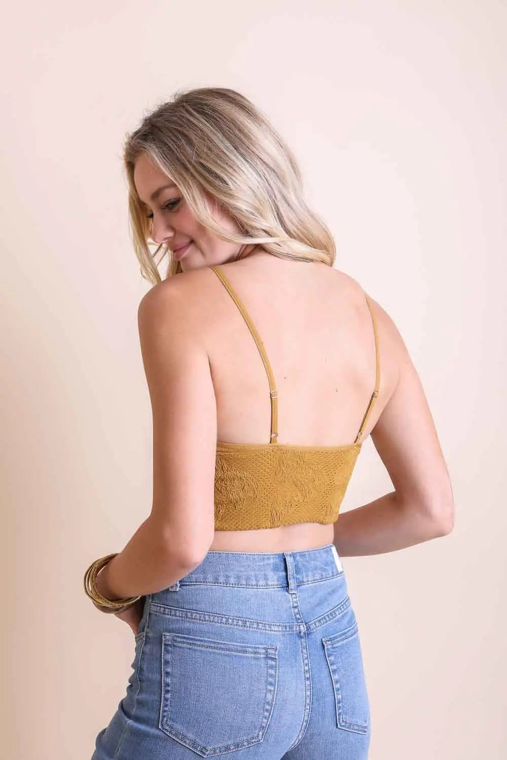 Rear view of woman modeling mustard yellow textured seamless brami with adjustable straps paired with light blue jeans against plain backdrop