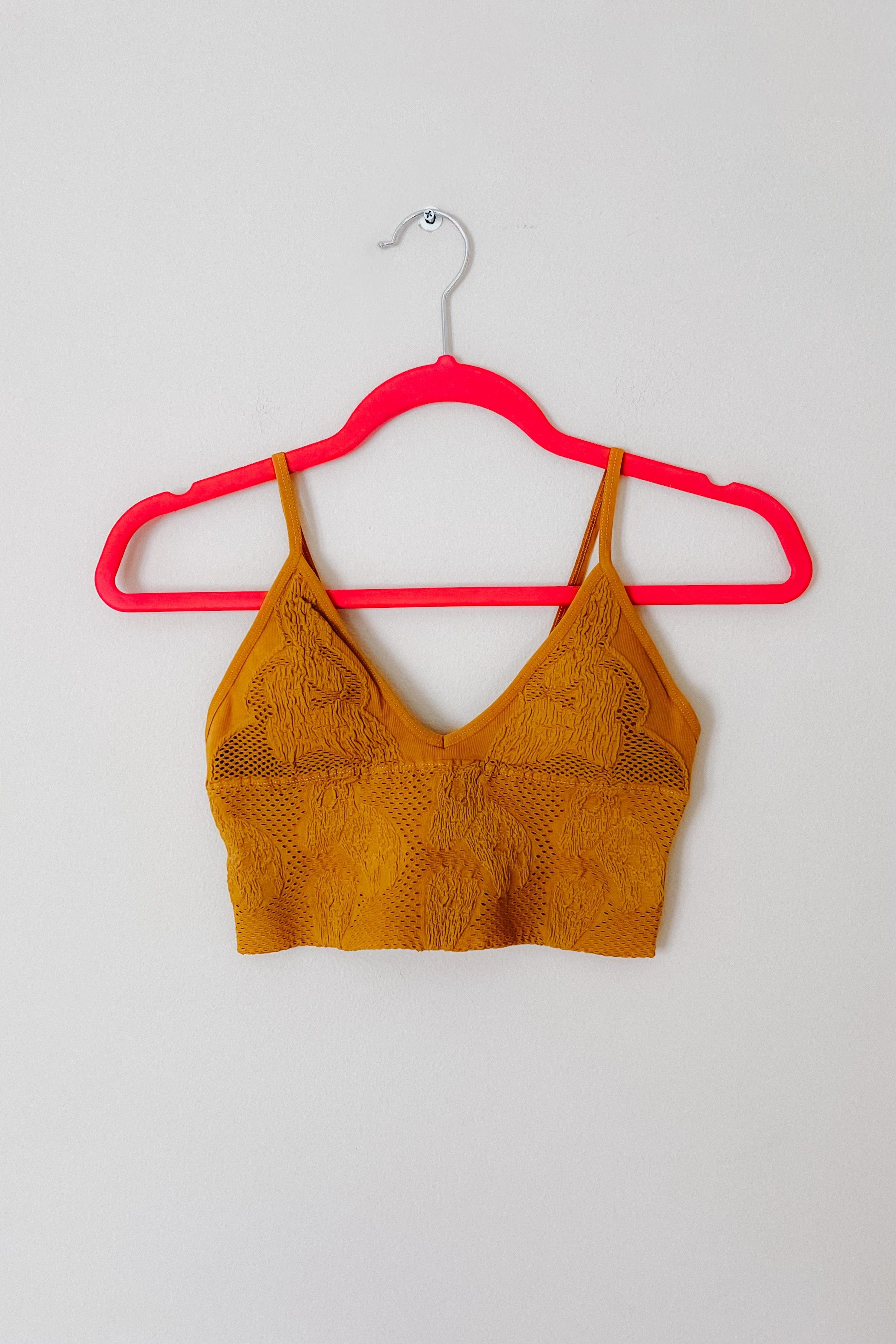 Mustard textured brami with intricate floral lace design and thin adjustable straps displayed on bright pink hanger against neutral wall