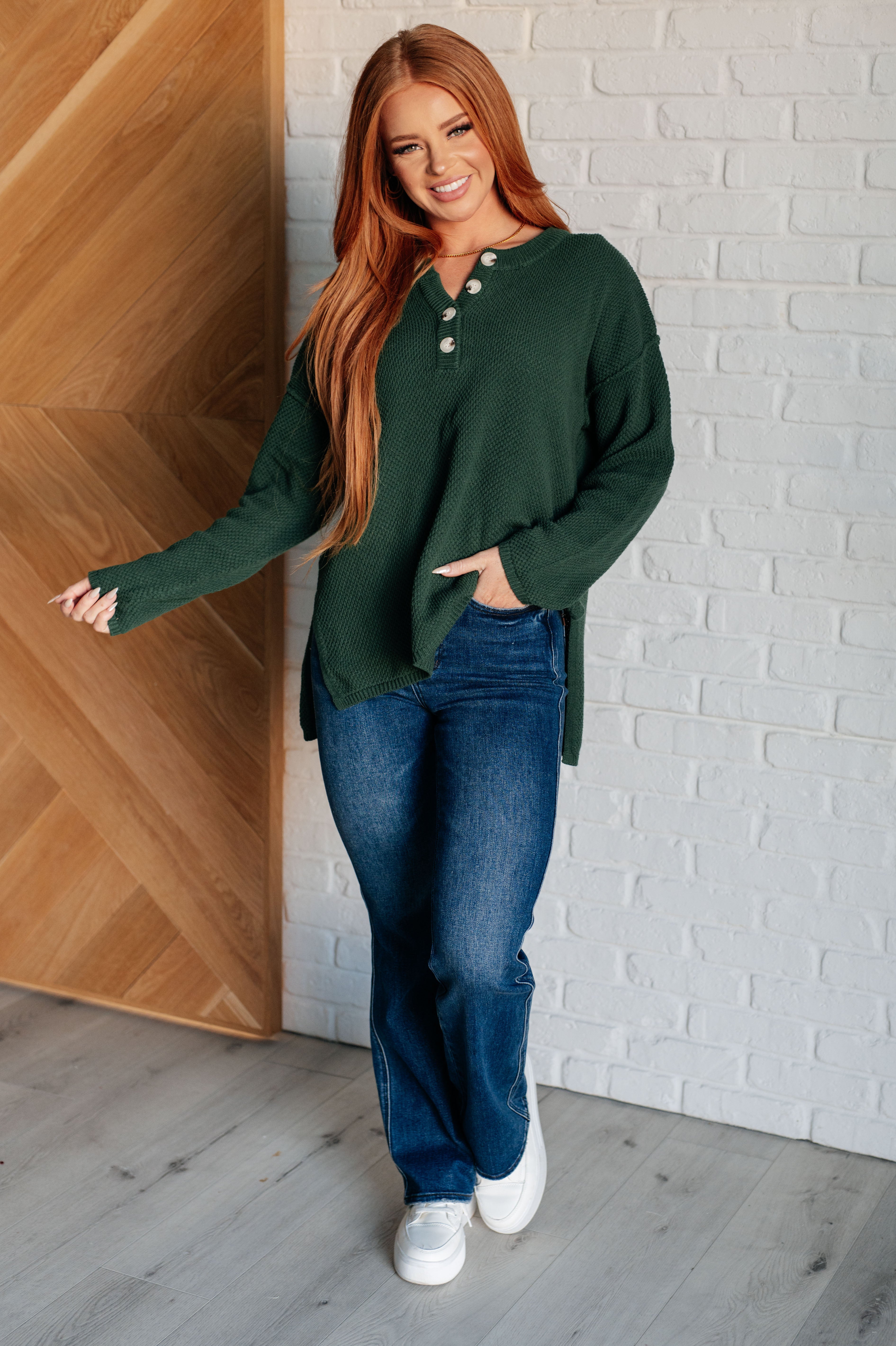 Young woman with long red hair wearing dark green waffle knit henley sweater and blue jeans standing indoors