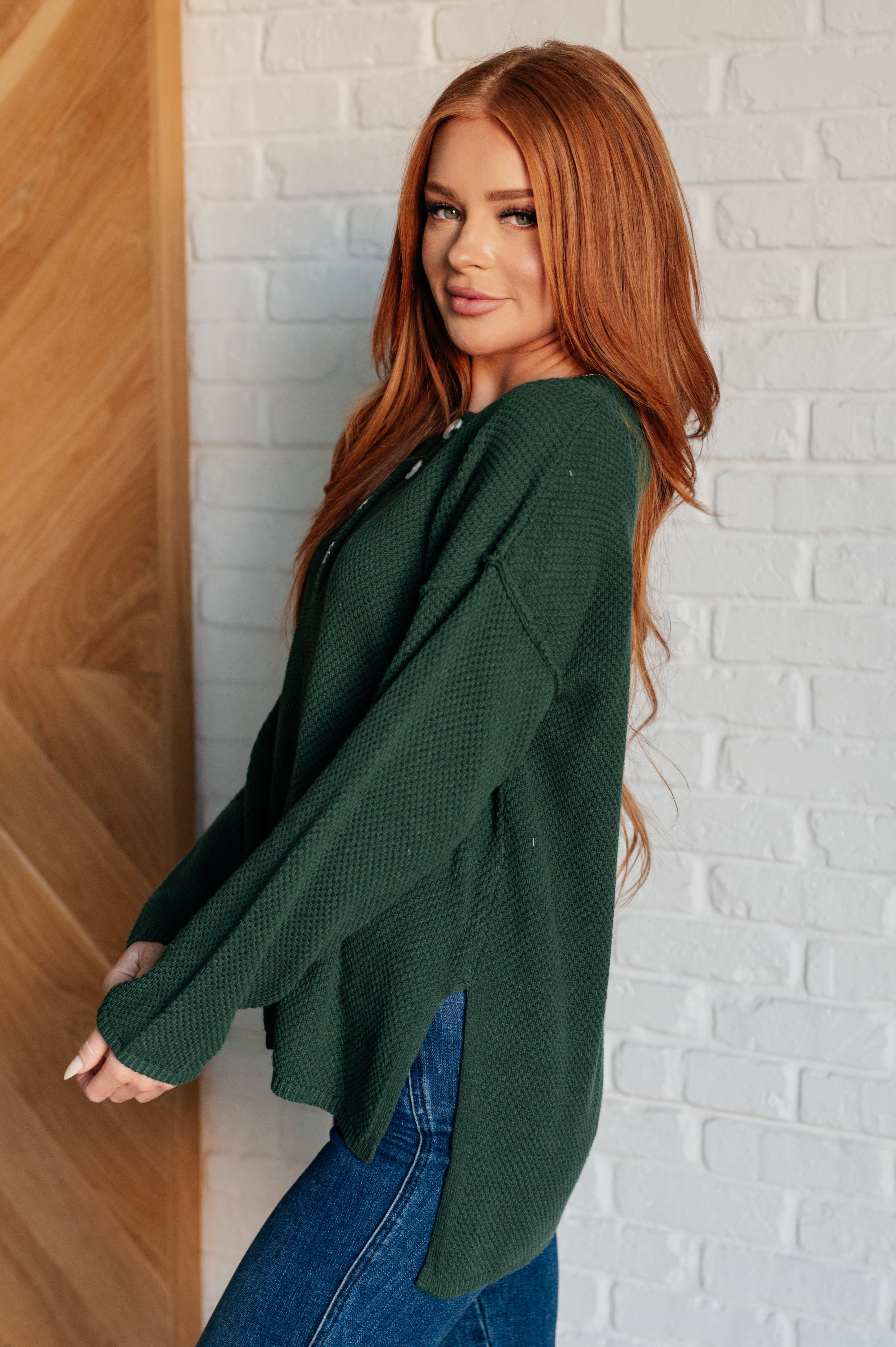Side profile of woman wearing dark green textured henley sweater with long sleeves and blue jeans indoors