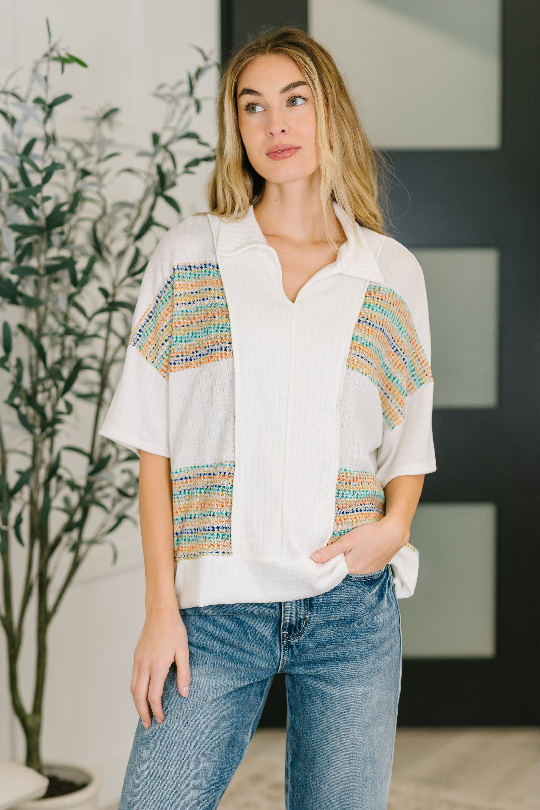 Woman wearing a cream knit top with colorful panels and a collared V-neck, paired with casual blue jeans in an eclectic space