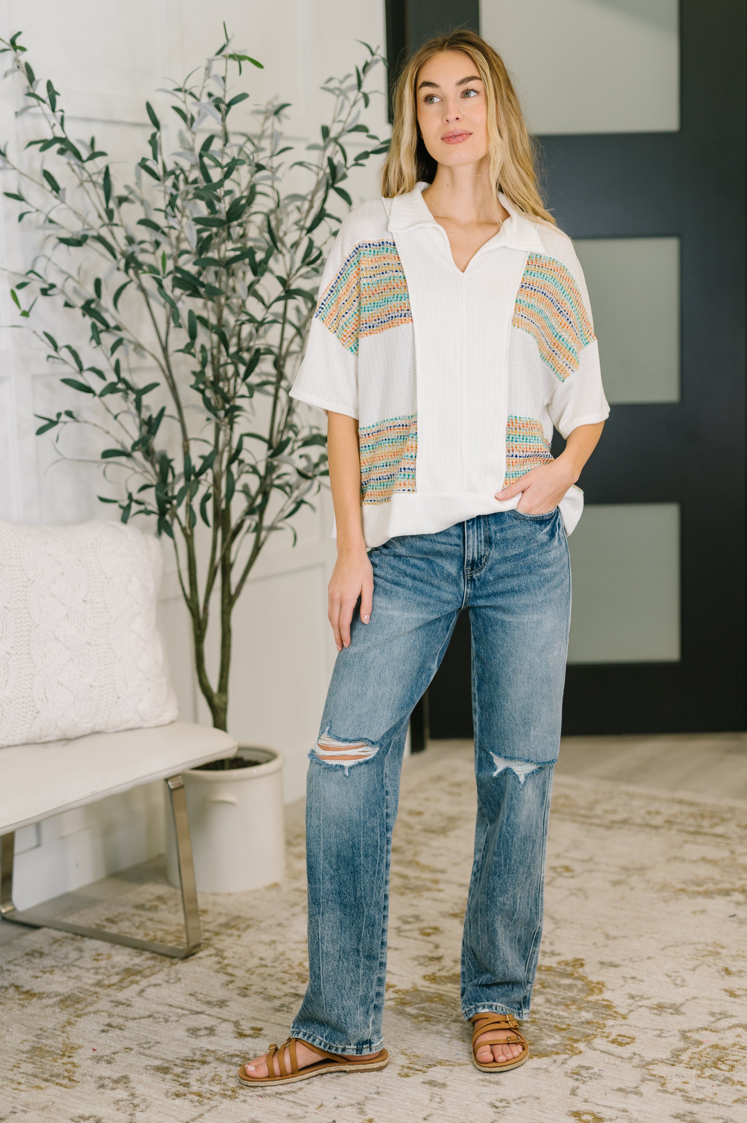 Woman wearing a cream collared knit top with colorful textured panels paired with distressed jeans in a relaxed eclectic room