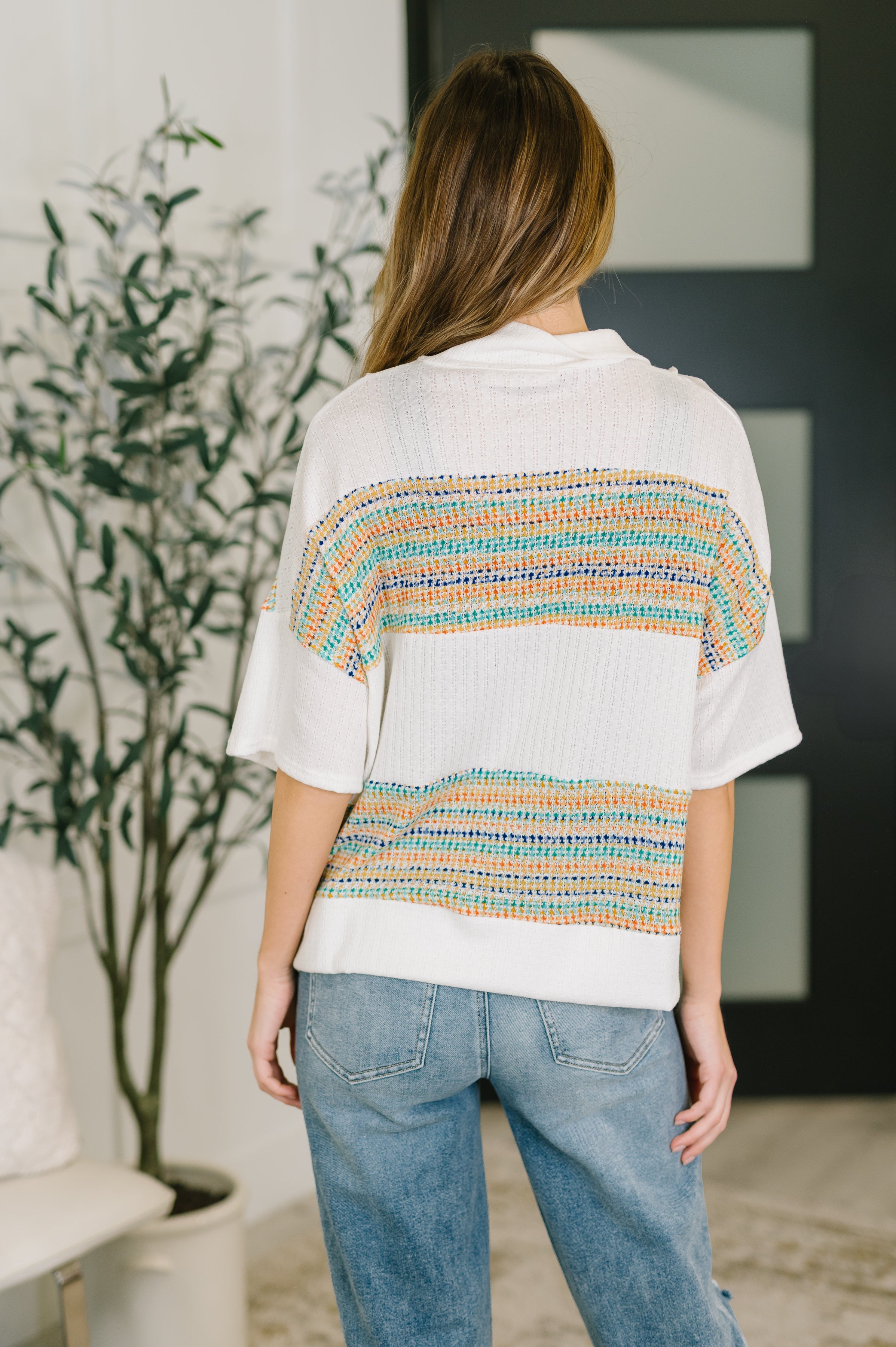 Woman wearing a cream knit top with colorful horizontal stripes and rolled sleeves in a cozy eclectic setting