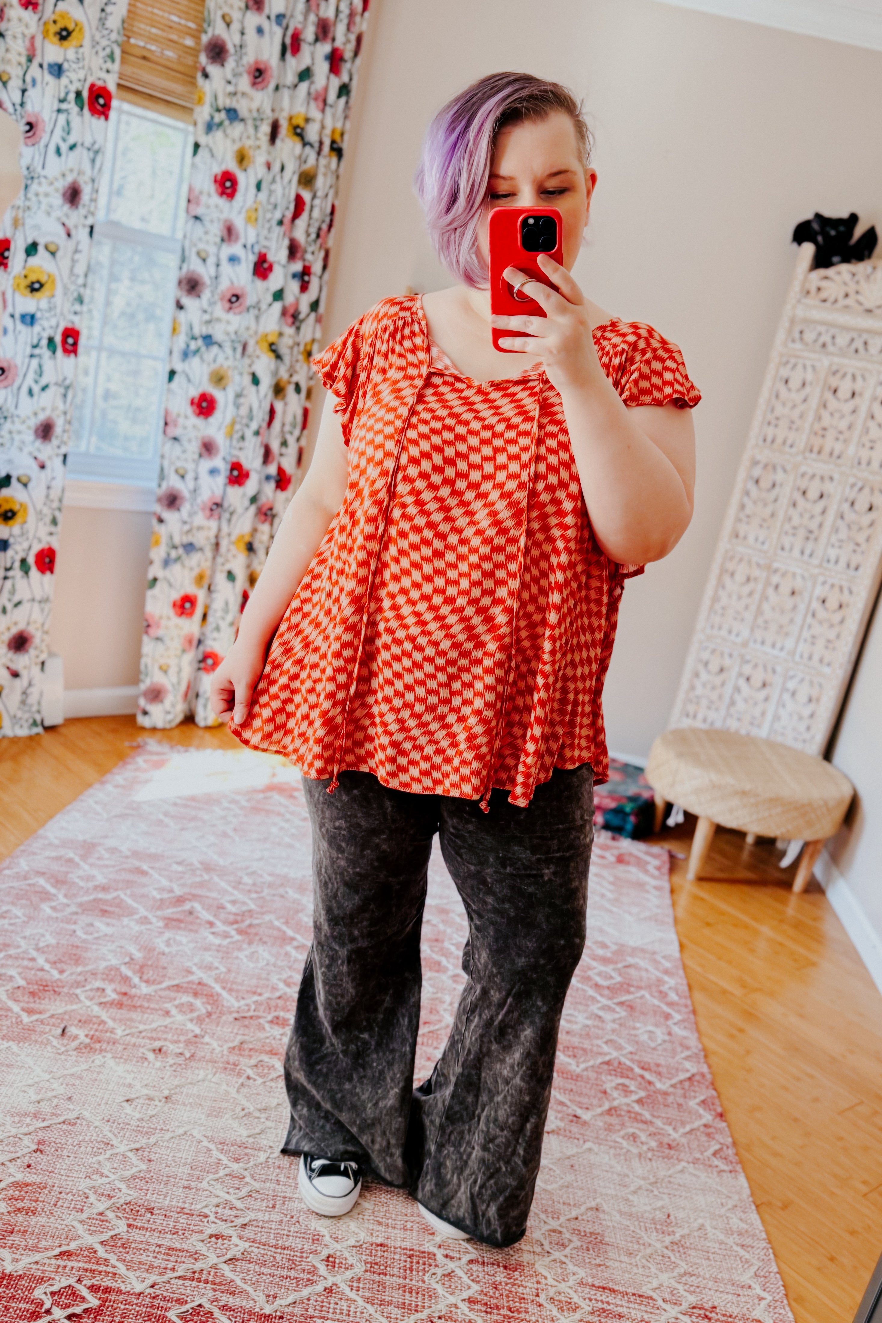 Woman with short purple hair wearing orange houndstooth butterfly sleeve top and black washed flare pants taking indoor mirror selfie