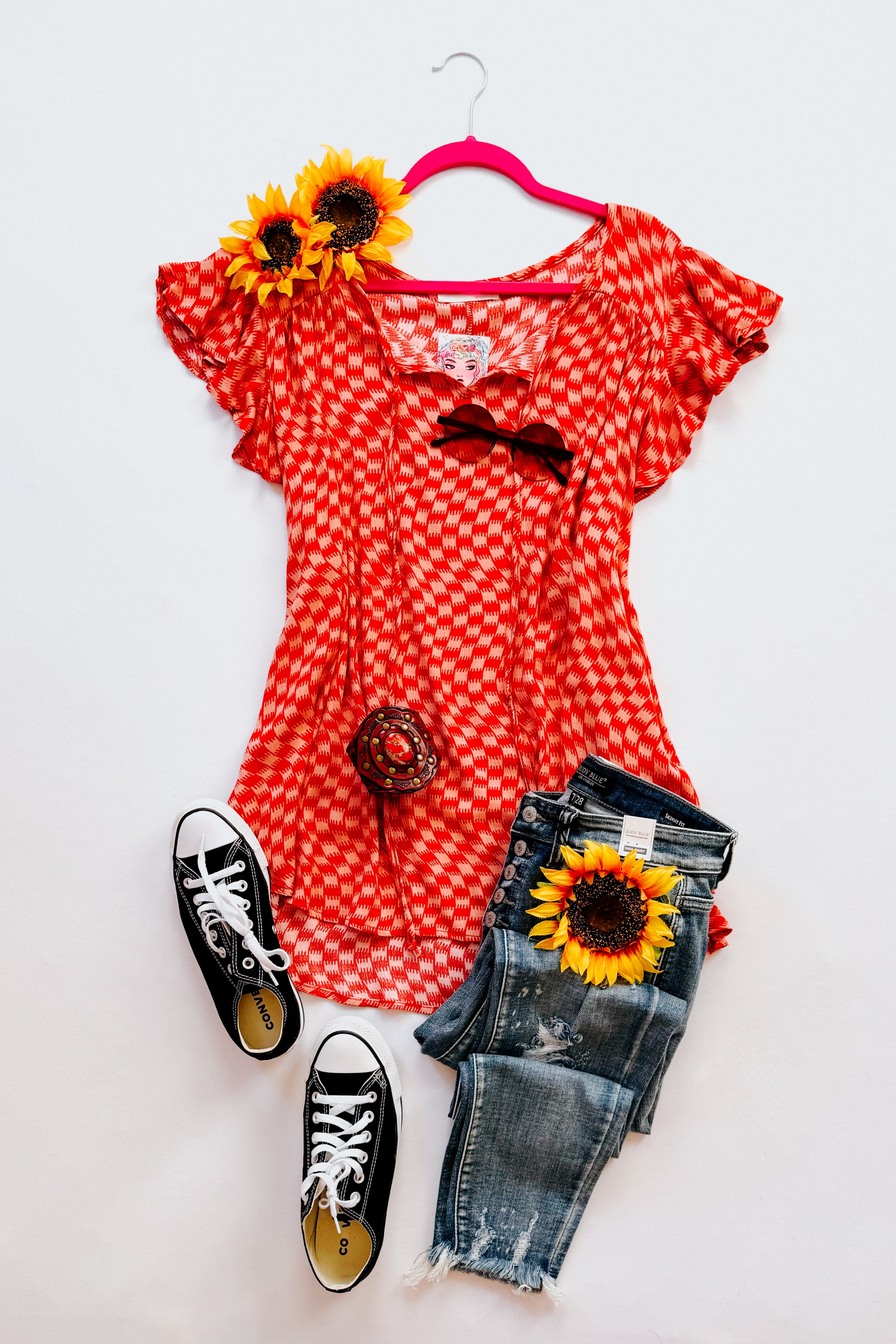 Red-orange houndstooth butterfly sleeve top styled with distressed jeans, black sneakers, sunglasses, and sunflowers on pink hanger