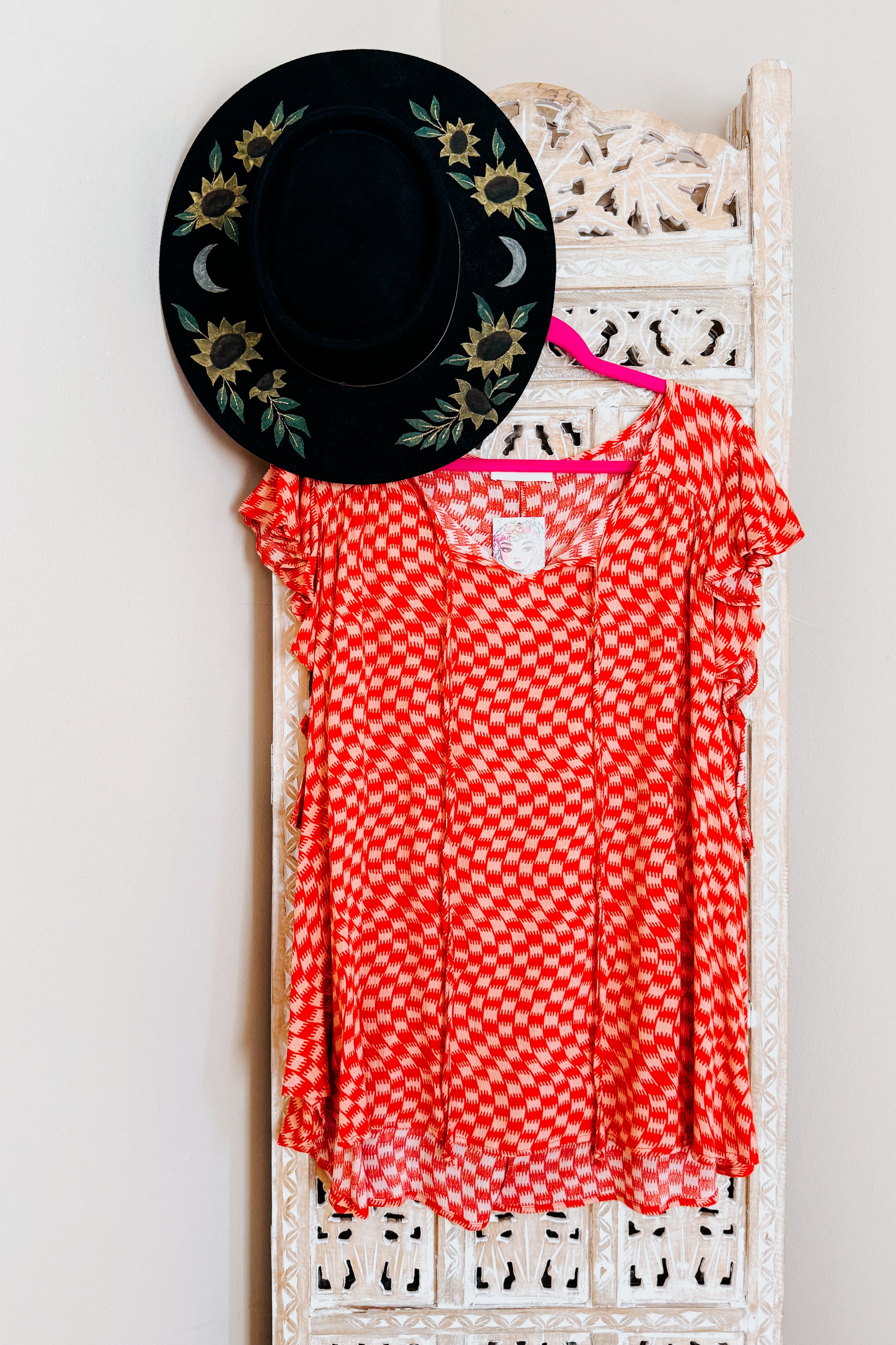 Orange houndstooth top with flutter sleeves displayed on pink hanger against carved white wooden screen with black embroidered hat