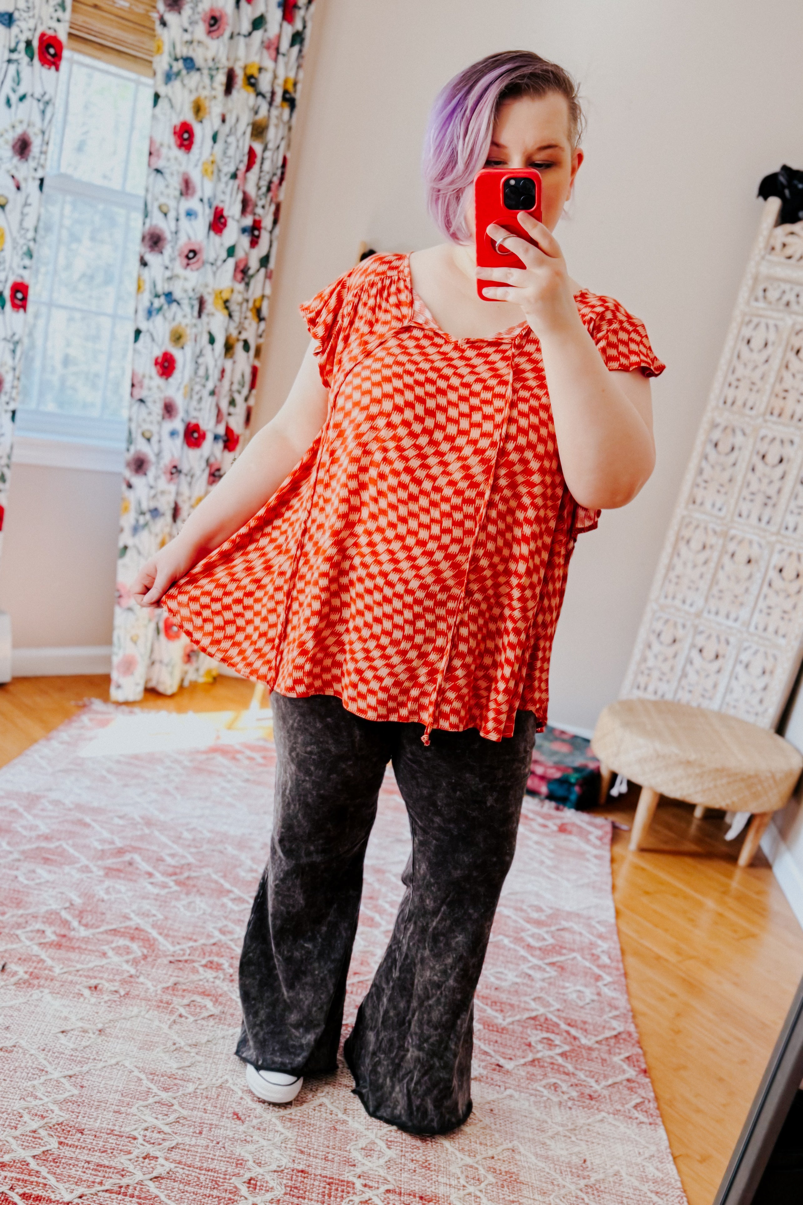 Woman with short purple hair wearing orange houndstooth butterfly sleeve top and black pants taking mirror selfie indoors