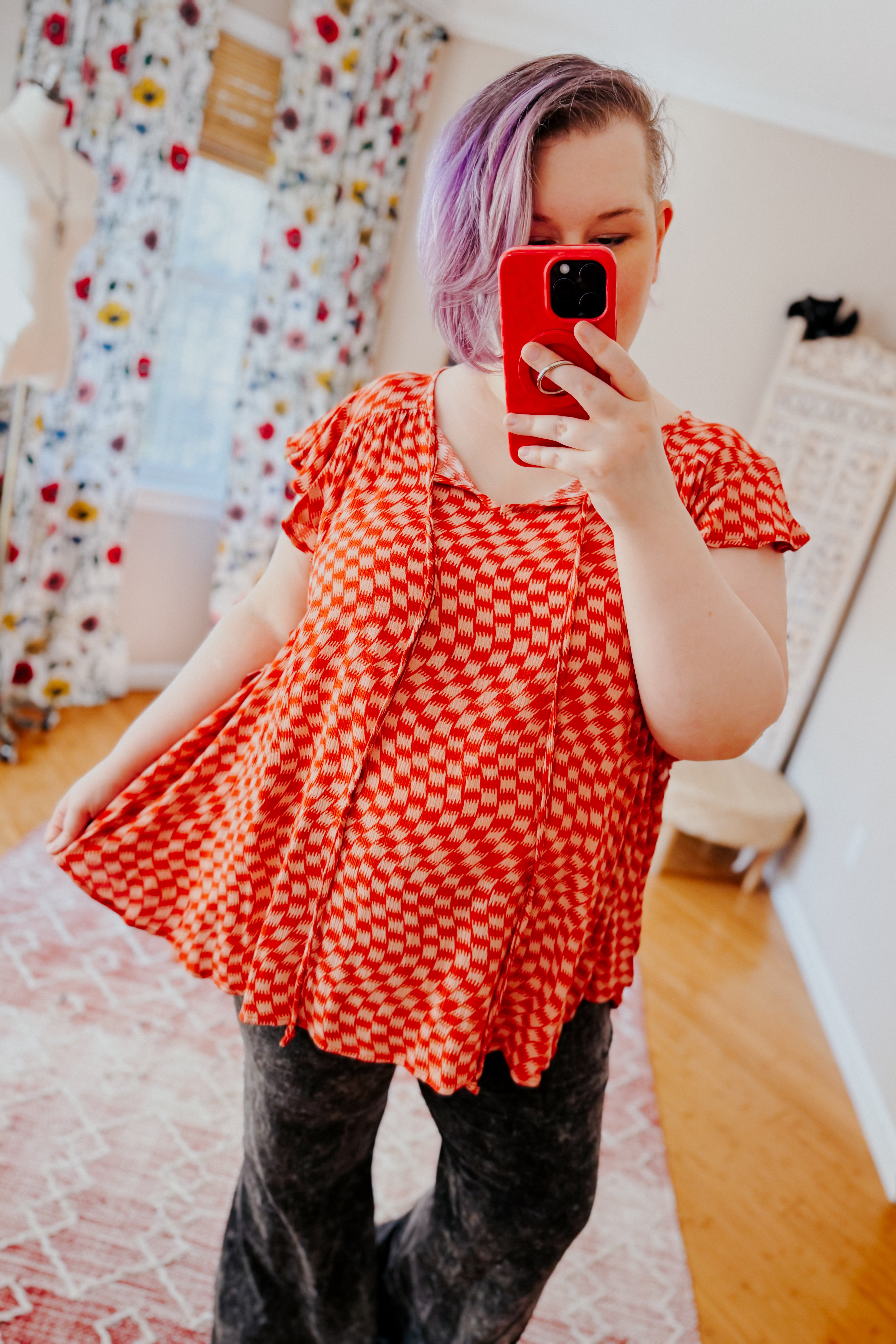 Woman with purple hair wearing orange houndstooth butterfly sleeve top taking mirror selfie indoors