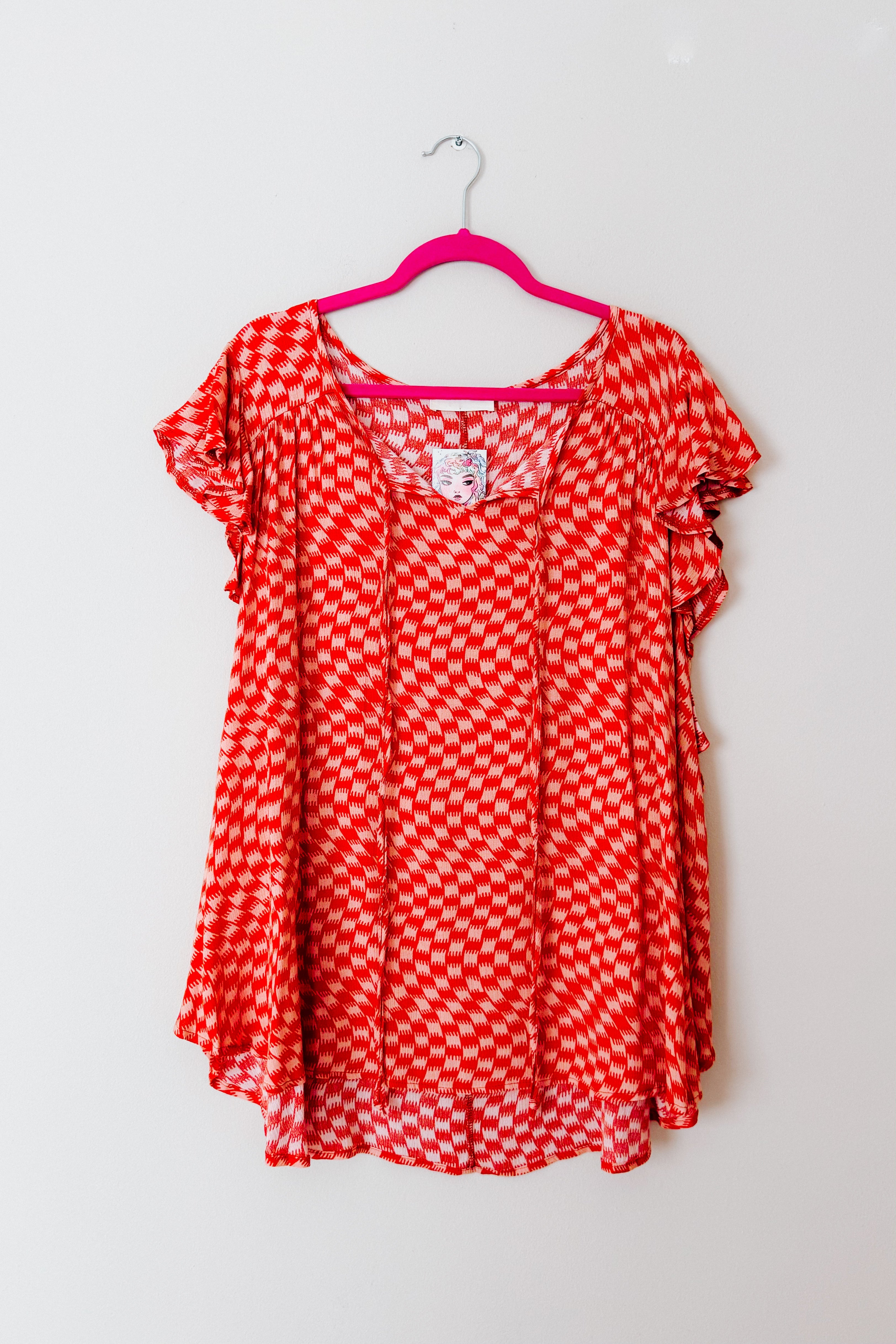 Orange houndstooth patterned top with flutter butterfly sleeves hanging on bright pink hanger against white wall
