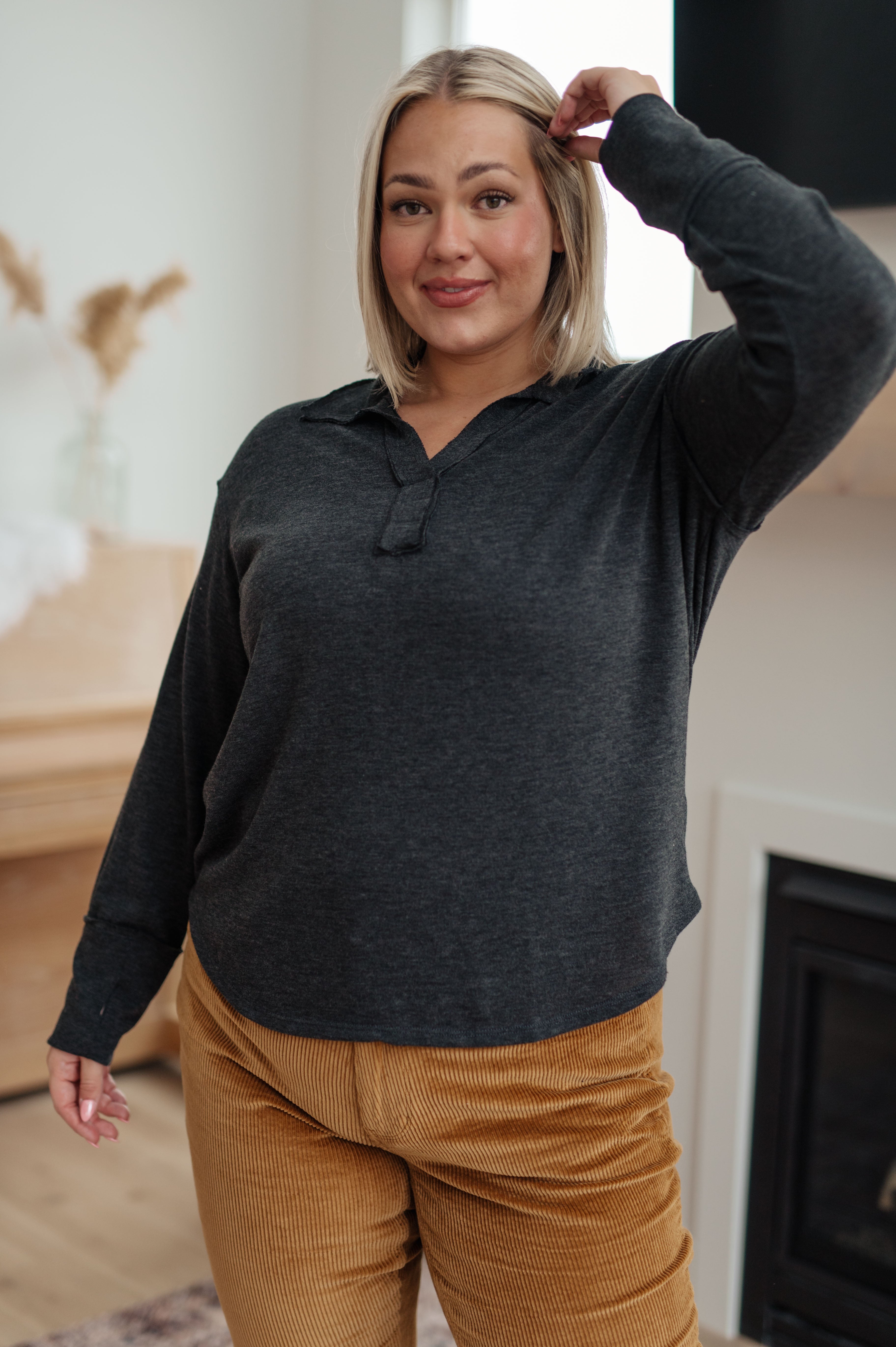 Smiling woman indoors wearing charcoal long sleeve Good Luck Charm top paired with mustard corduroy pants in warm living space