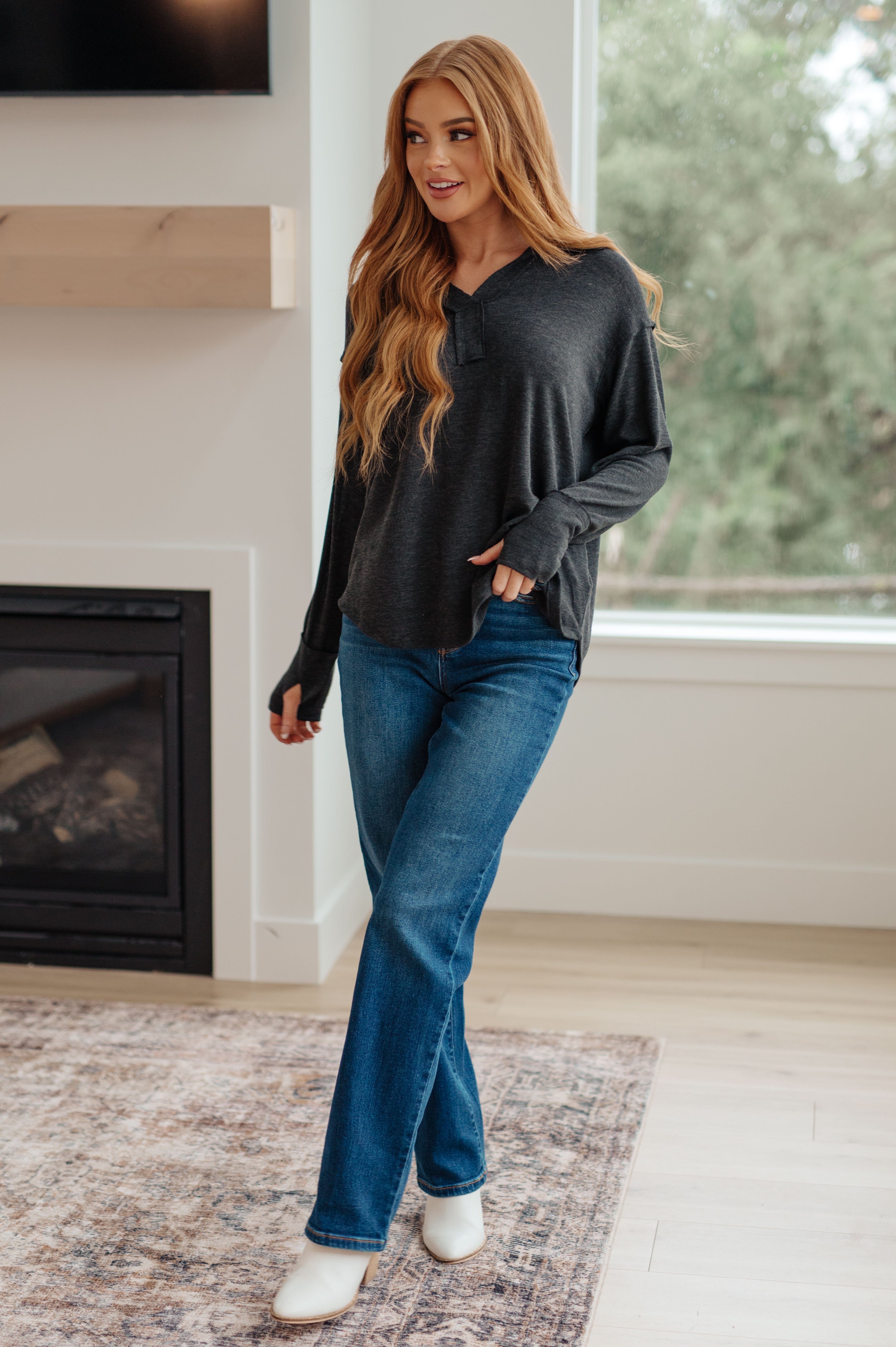 Woman wearing charcoal long sleeve Good Luck Charm top paired with blue jeans and white boots in bright room with fireplace and window