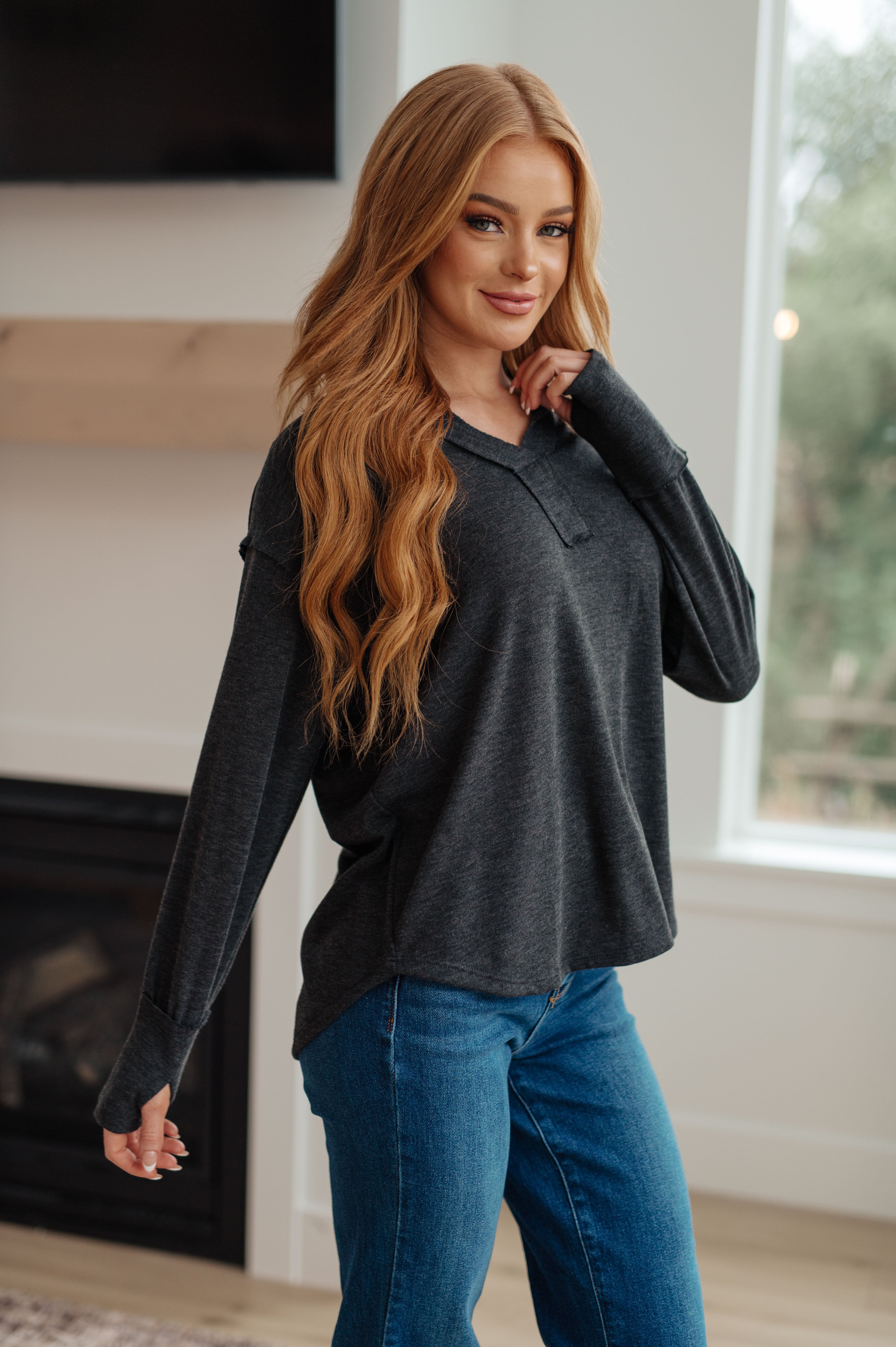 Woman standing indoors wearing charcoal long sleeve Good Luck Charm top paired with blue jeans near window