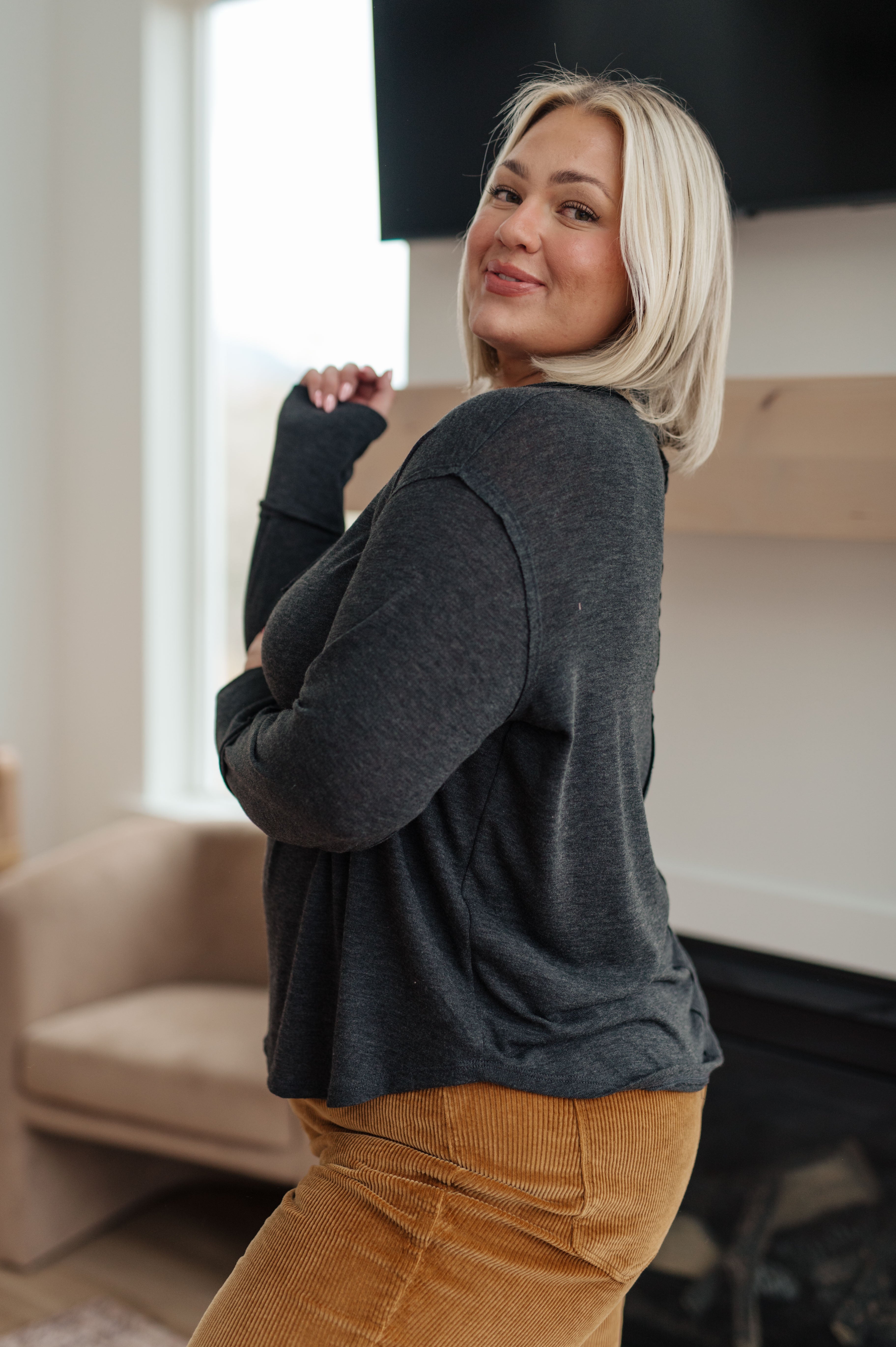 Smiling woman with blonde hair wearing charcoal long sleeve top and mustard corduroy pants in cozy living room setting
