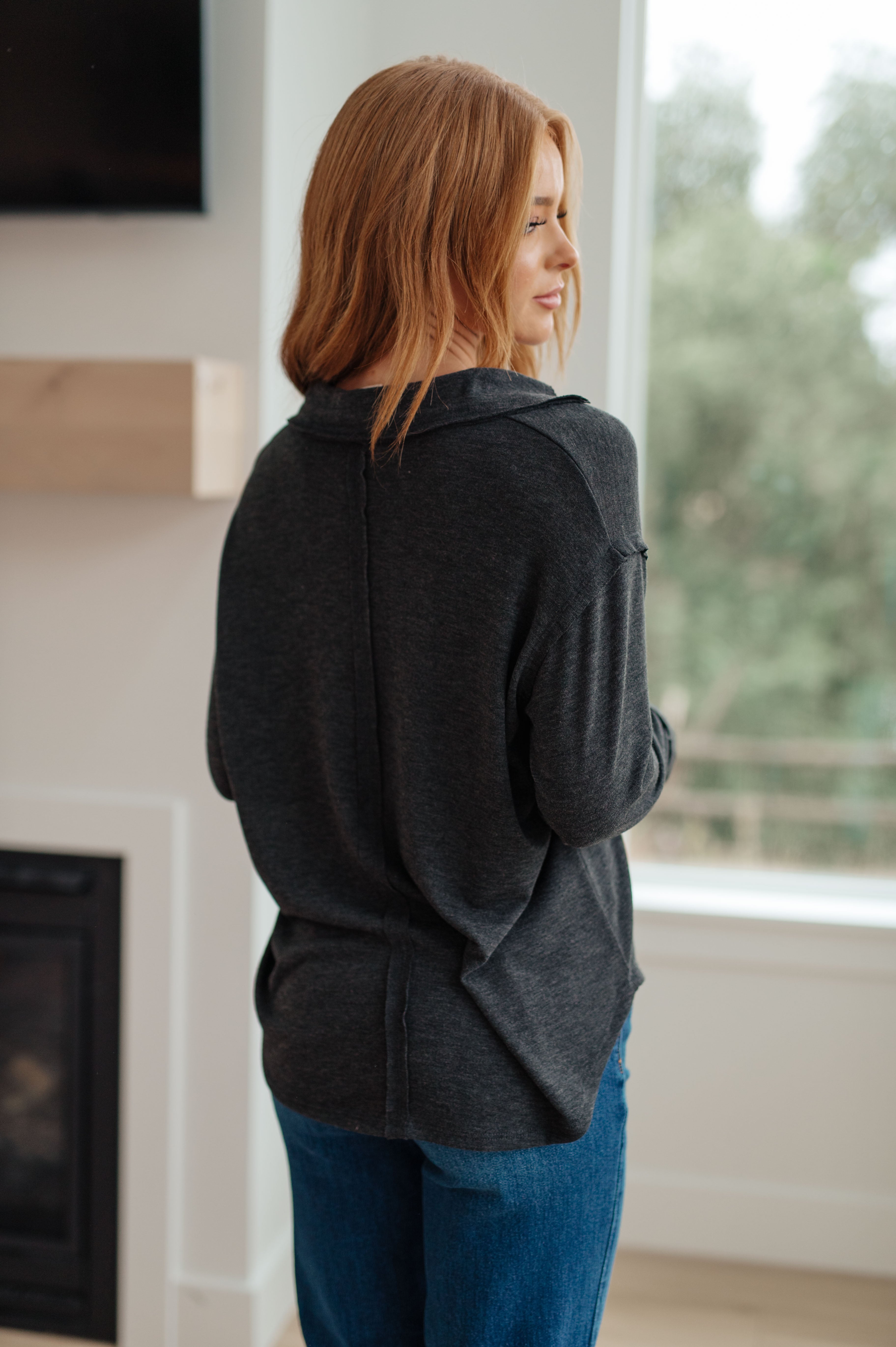 Woman seen from behind wearing charcoal long sleeve top with subtle seam details paired with blue jeans near window