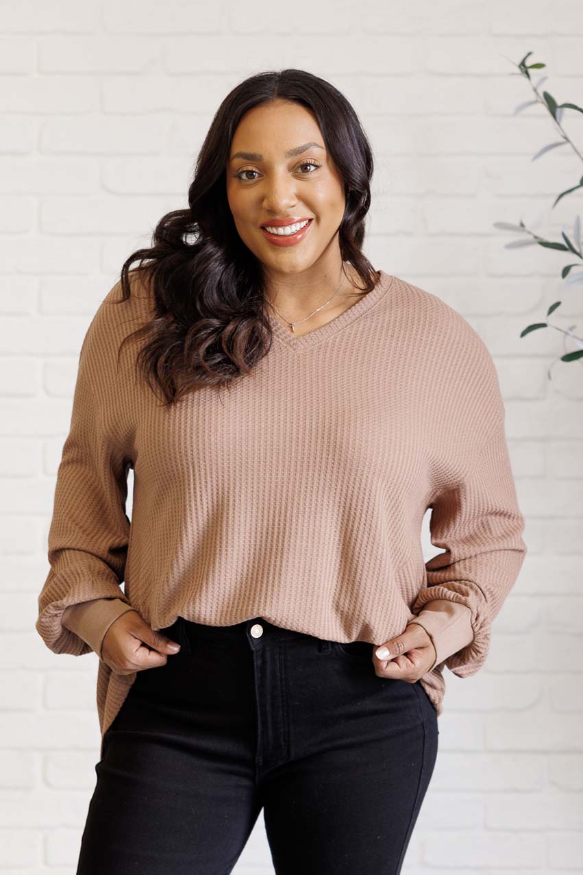 Smiling woman wearing a coffee V-neck knit top with drop shoulders and asymmetrical hem with black jeans indoors