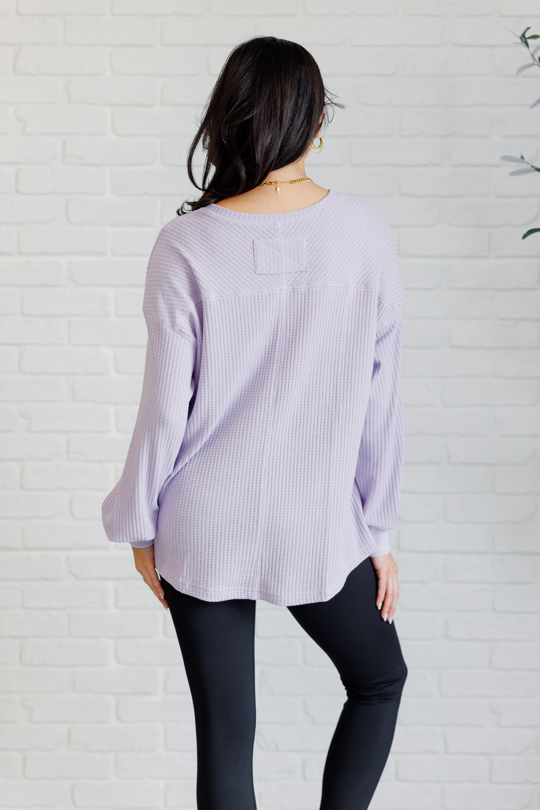 Woman wearing a lavender waffle knit long sleeve top and black leggings standing against a white brick wall.