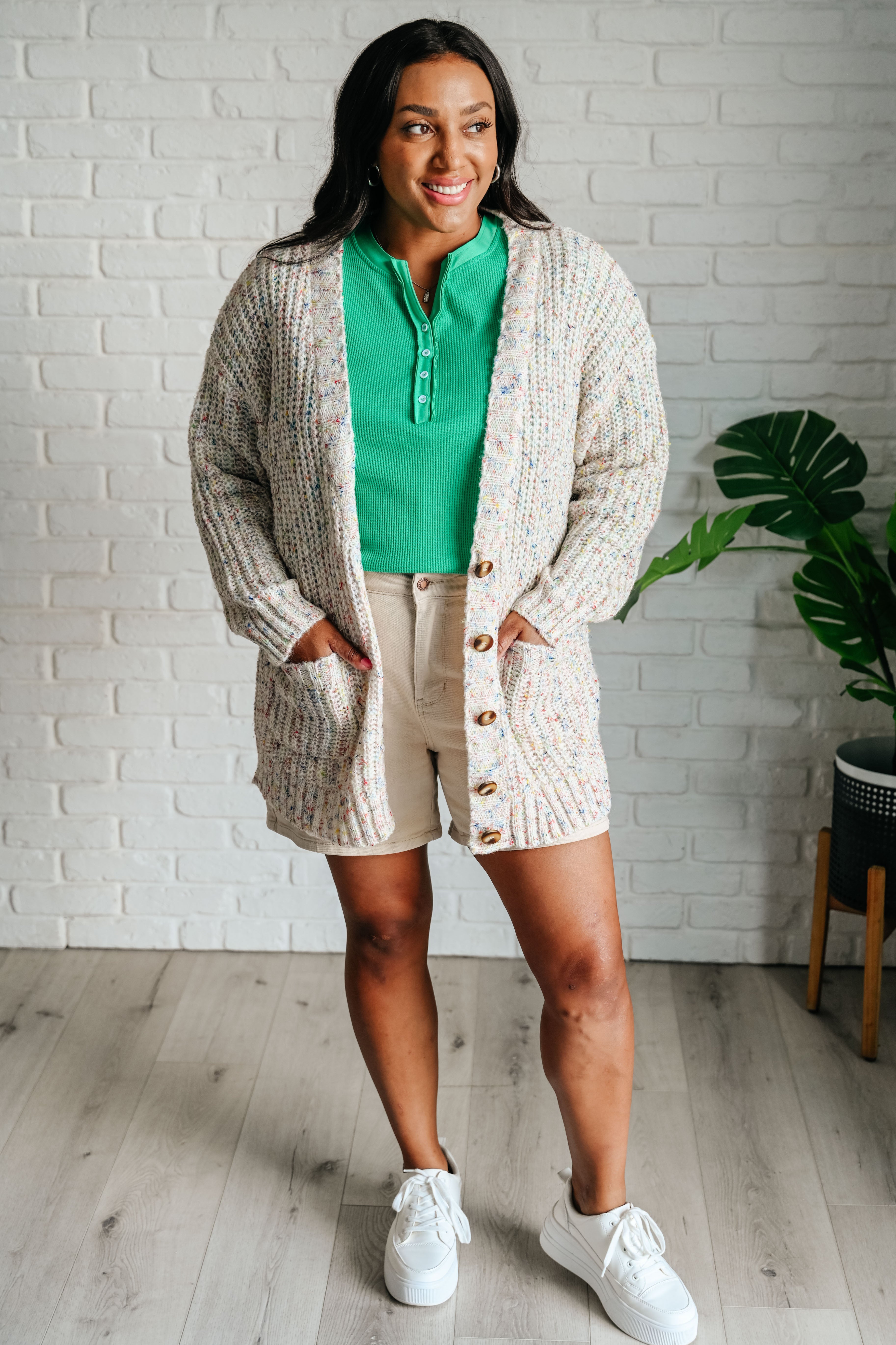 Woman wearing a green long-sleeve waffle knit henley top, a cardigan, beige shorts and white sneakers with a white brick wall background.