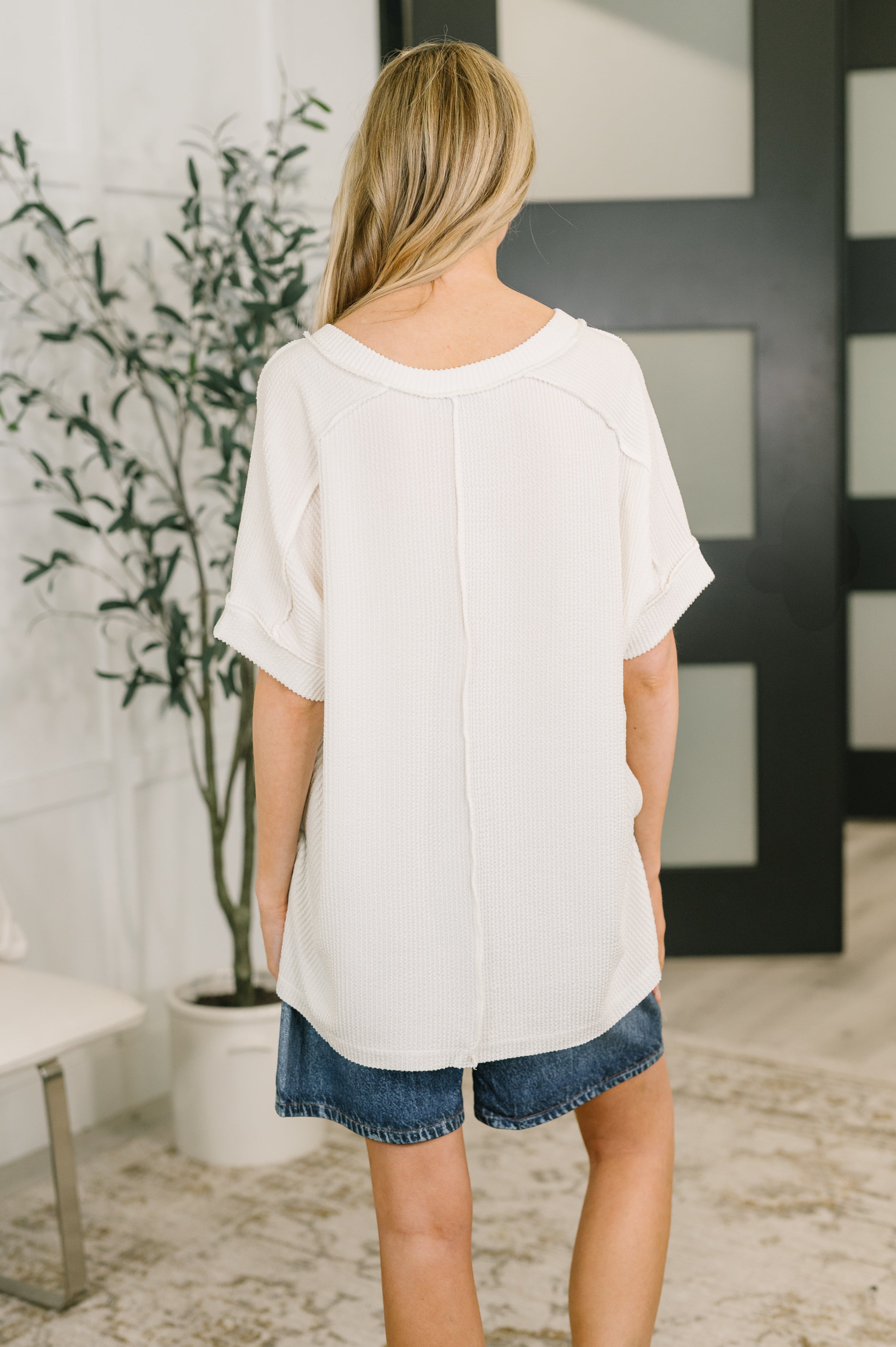 Woman indoors wearing a relaxed white textured short-sleeve top with a raw edge seam detail paired with denim shorts