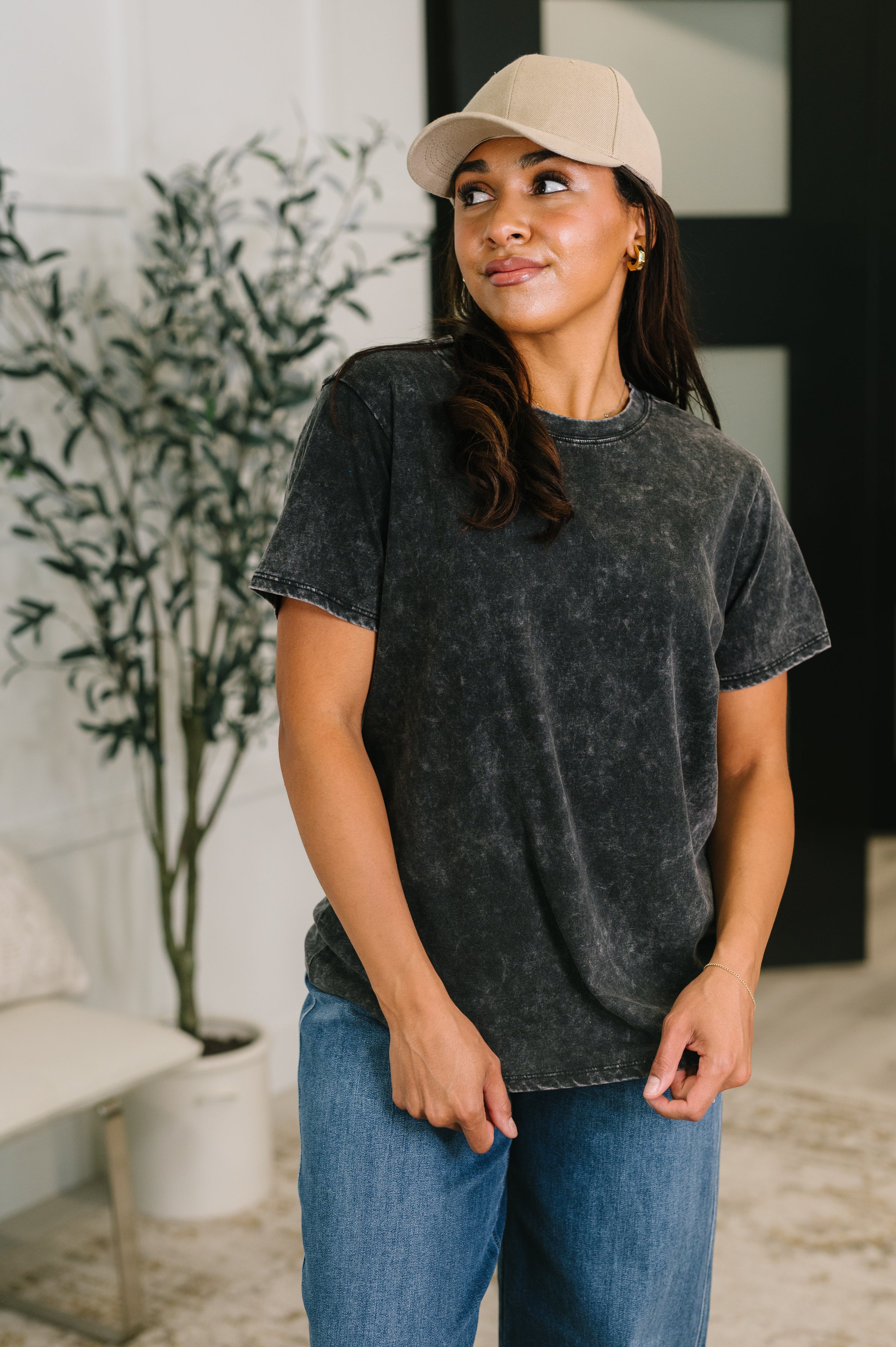 Woman wearing a mineral wash ash black tee with short sleeves paired with relaxed blue jeans and a beige cap indoors