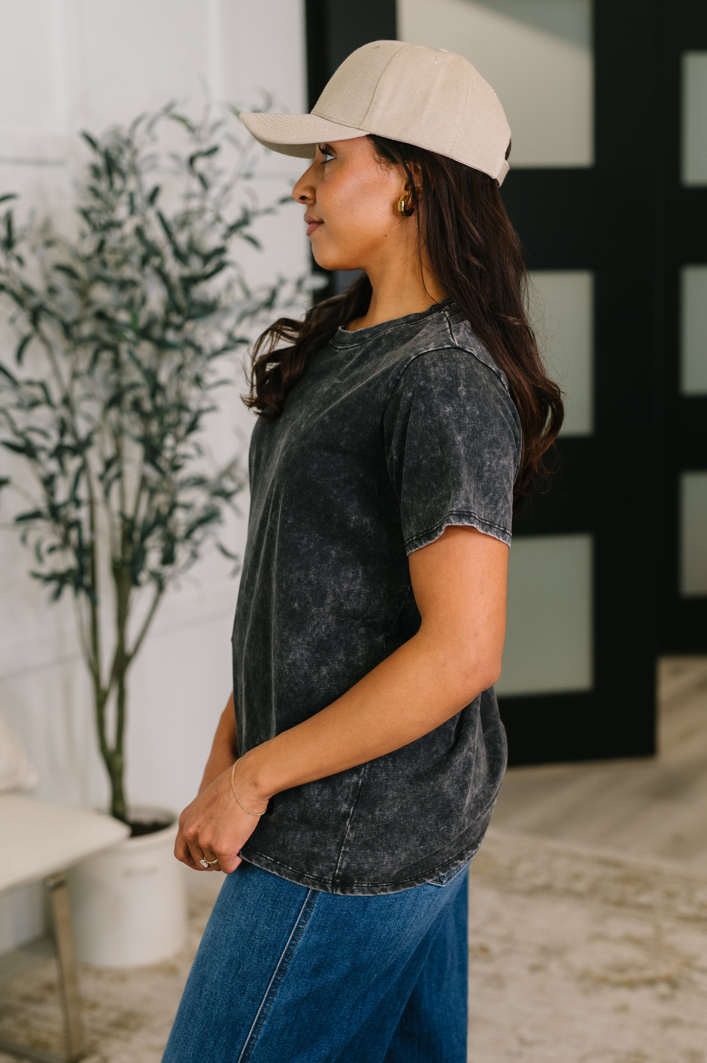 Profile of woman wearing ash black mineral wash tee paired with blue jeans and beige cap indoors
