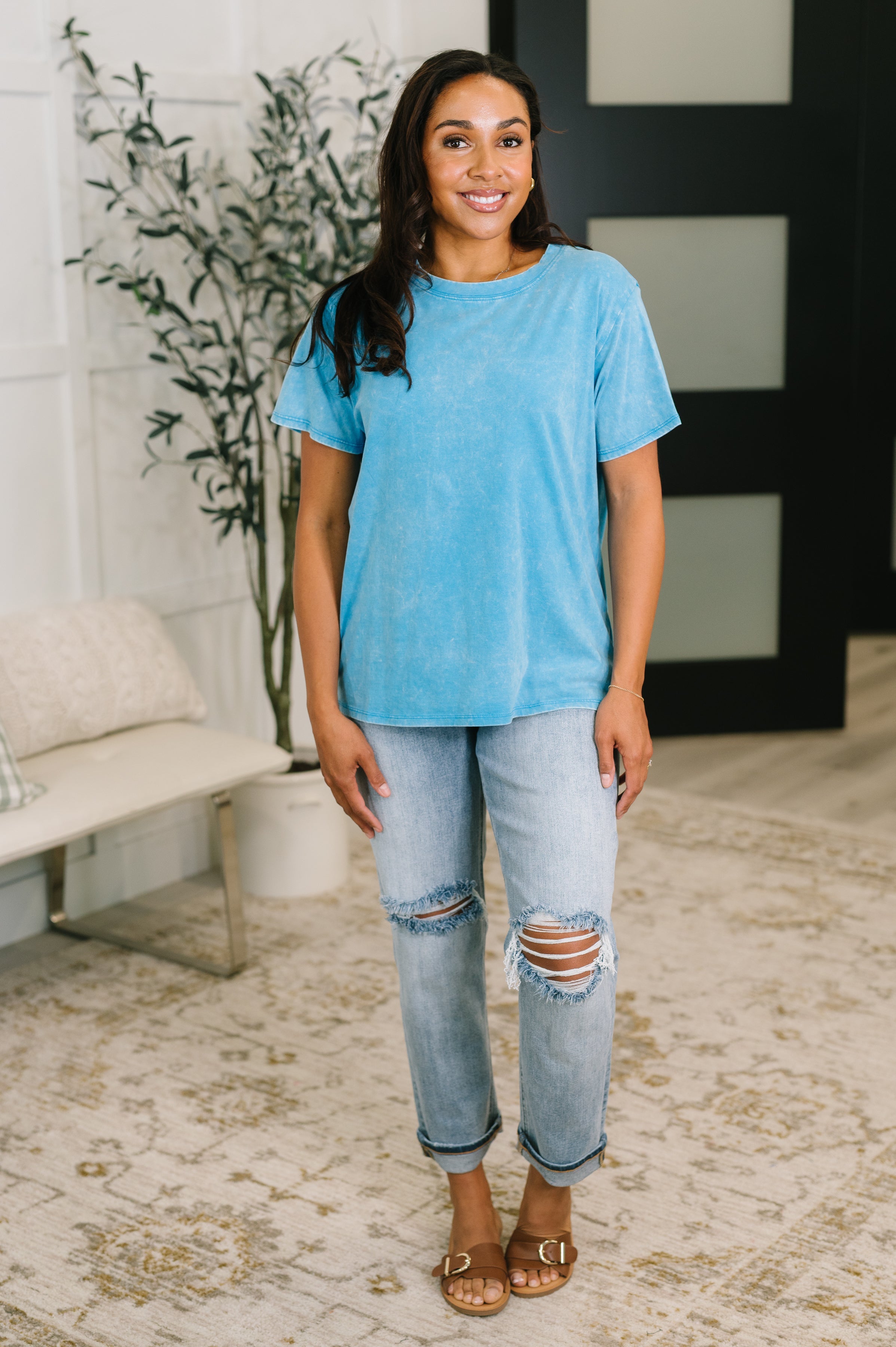 Woman standing indoors wearing a bold blue mineral wash tee with distressed light wash jeans and brown sandals