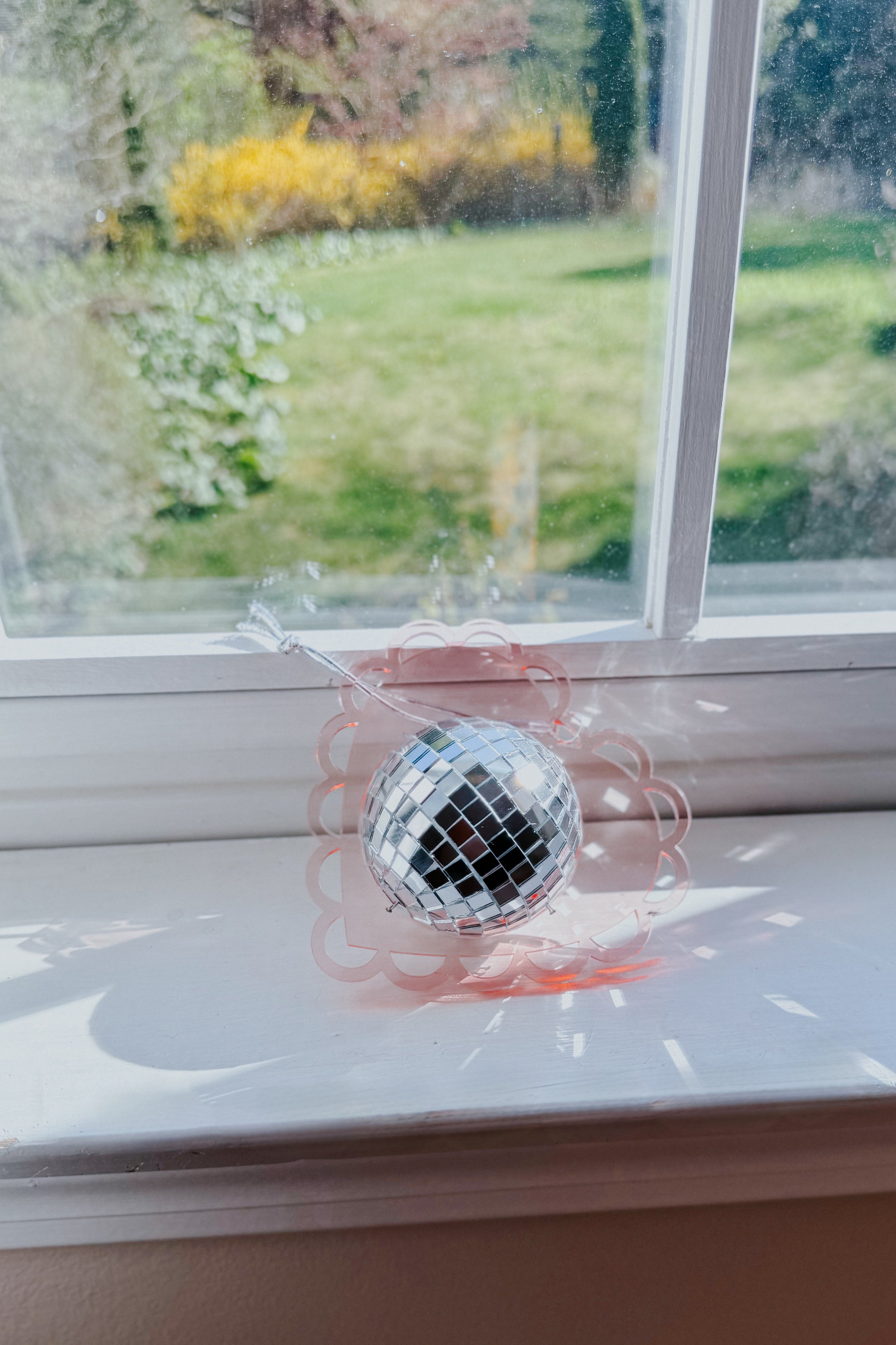 Soft pink scalloped heart with reflective silver mini disco ball resting on white windowsill with garden view behind