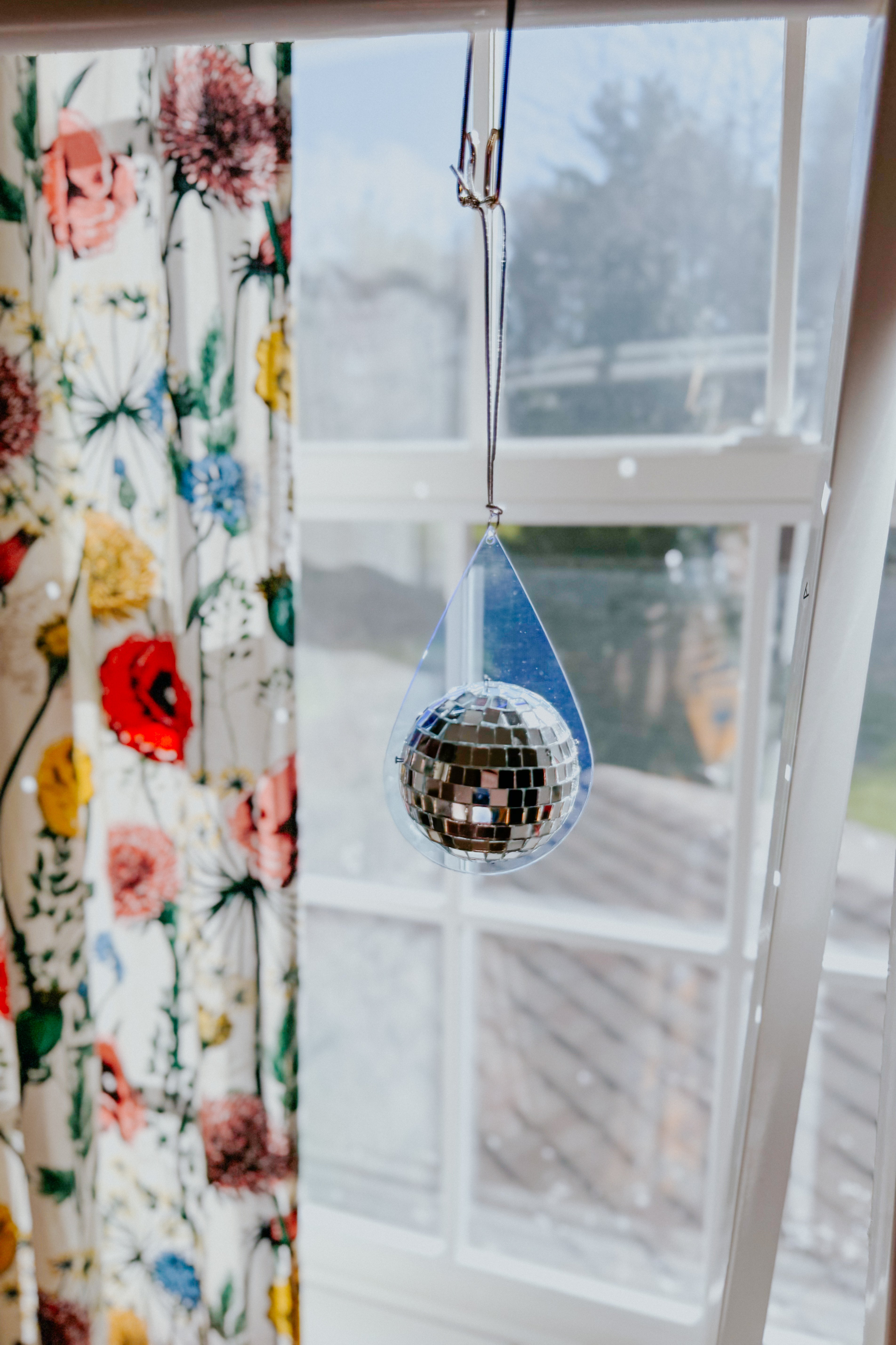 Light blue teardrop sun catcher with suspended mirrored mini disco ball hanging in front of window with floral curtain background