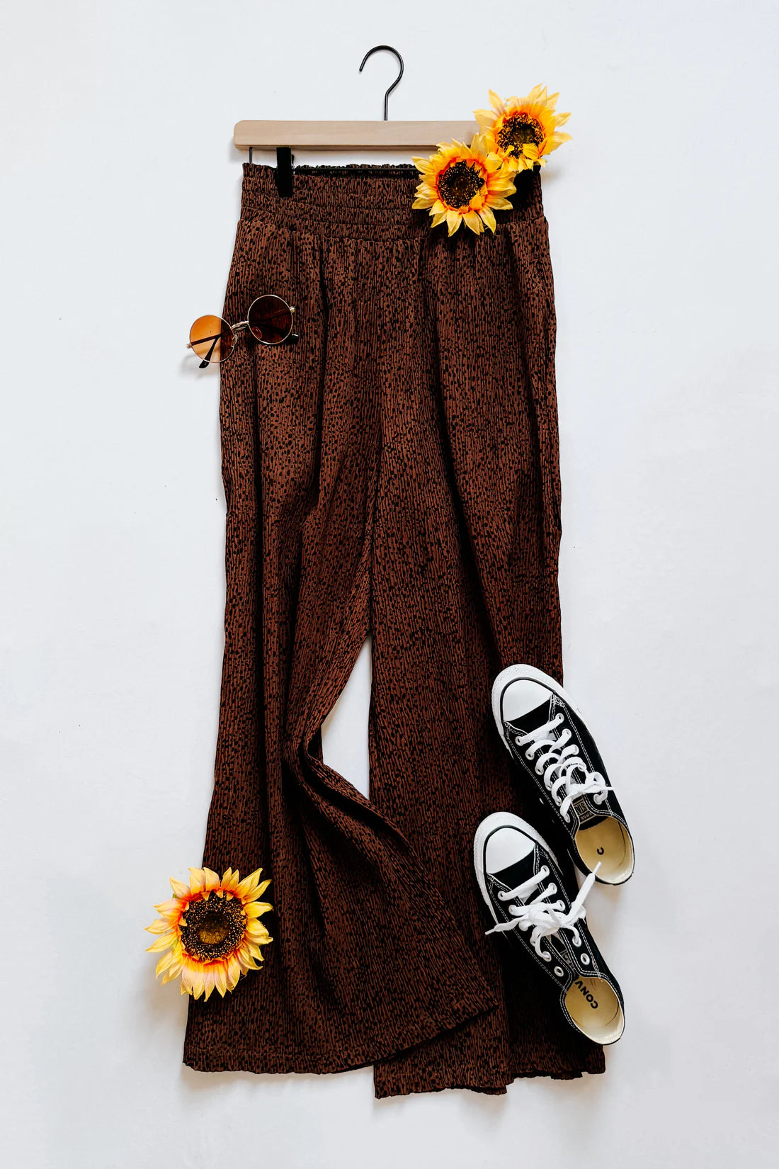 Brown high rise wide leg pants with subtle pattern styled with round sunglasses and black lace-up canvas shoes on white background