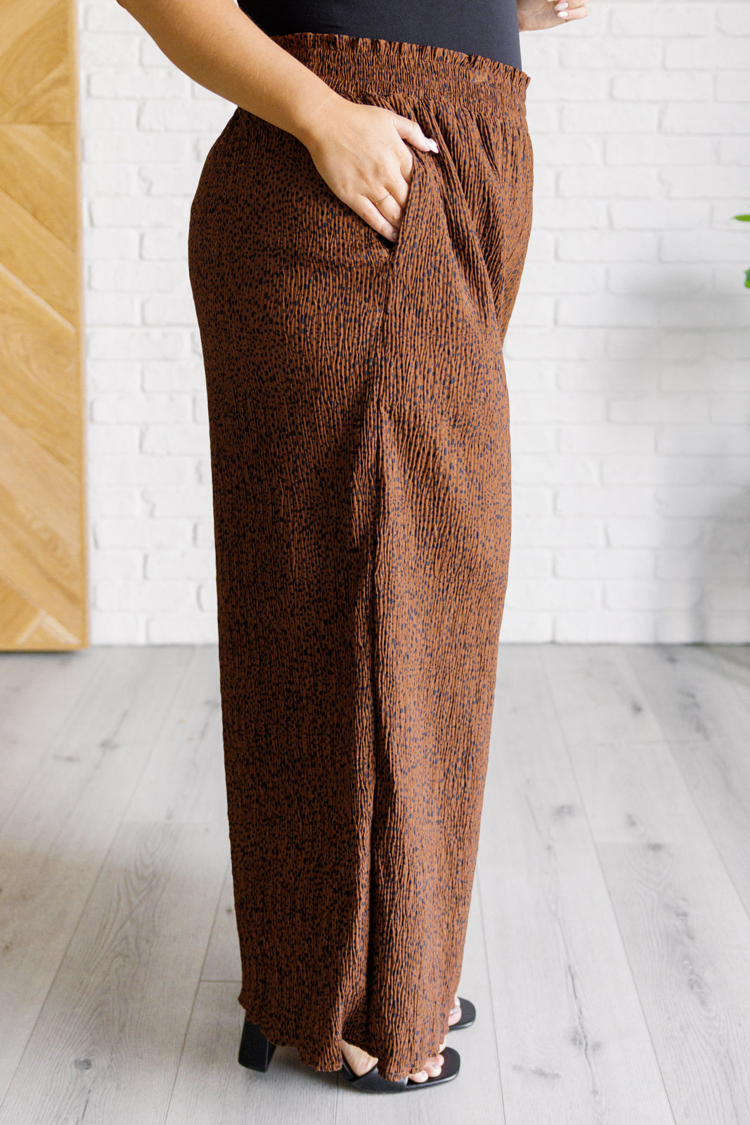Side profile of brown Harmony high rise wide leg pants with elastic waistband and subtle pattern worn with black top indoors